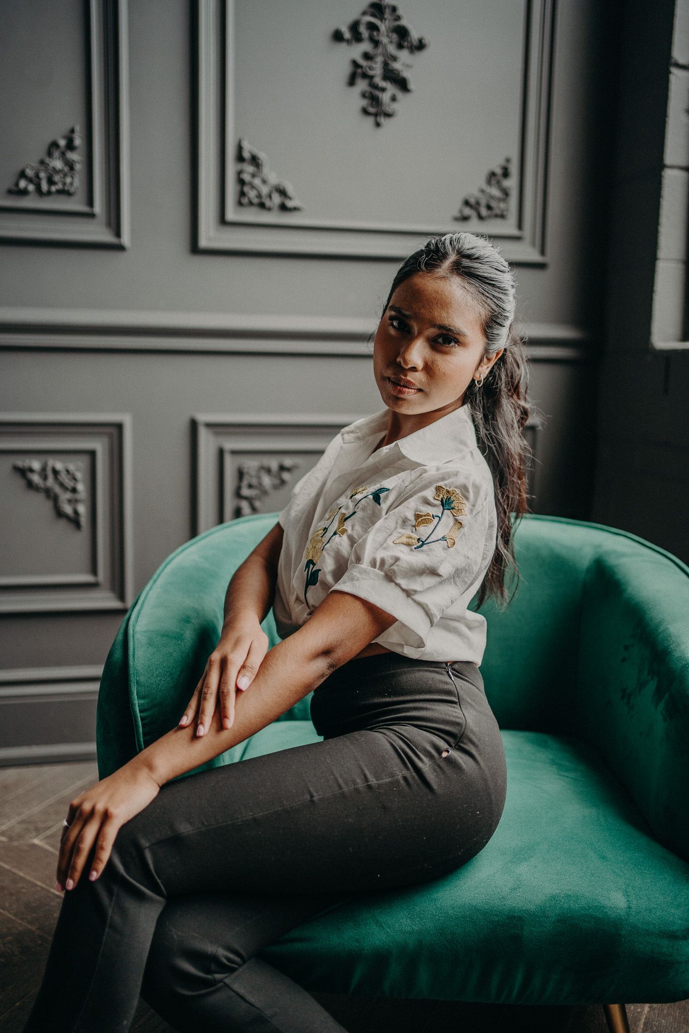 A young woman in a floral blouse and dark pants sits elegantly on a teal velvet armchair in a gray paneled room.