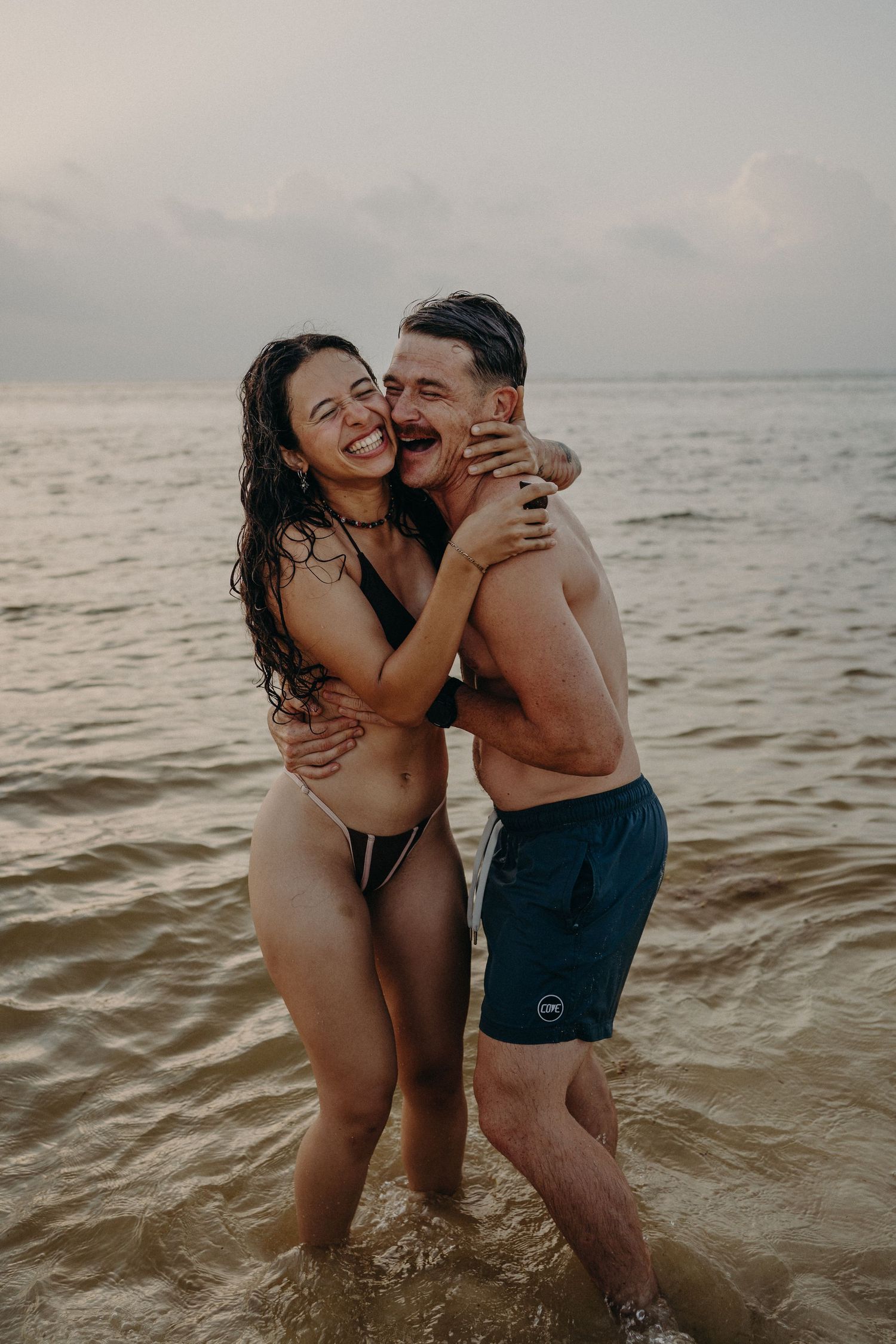 Laughing couple in swimwear embrace while standing in shallow ocean water at sunset, with calm waves and a soft cloudy sky behind them.