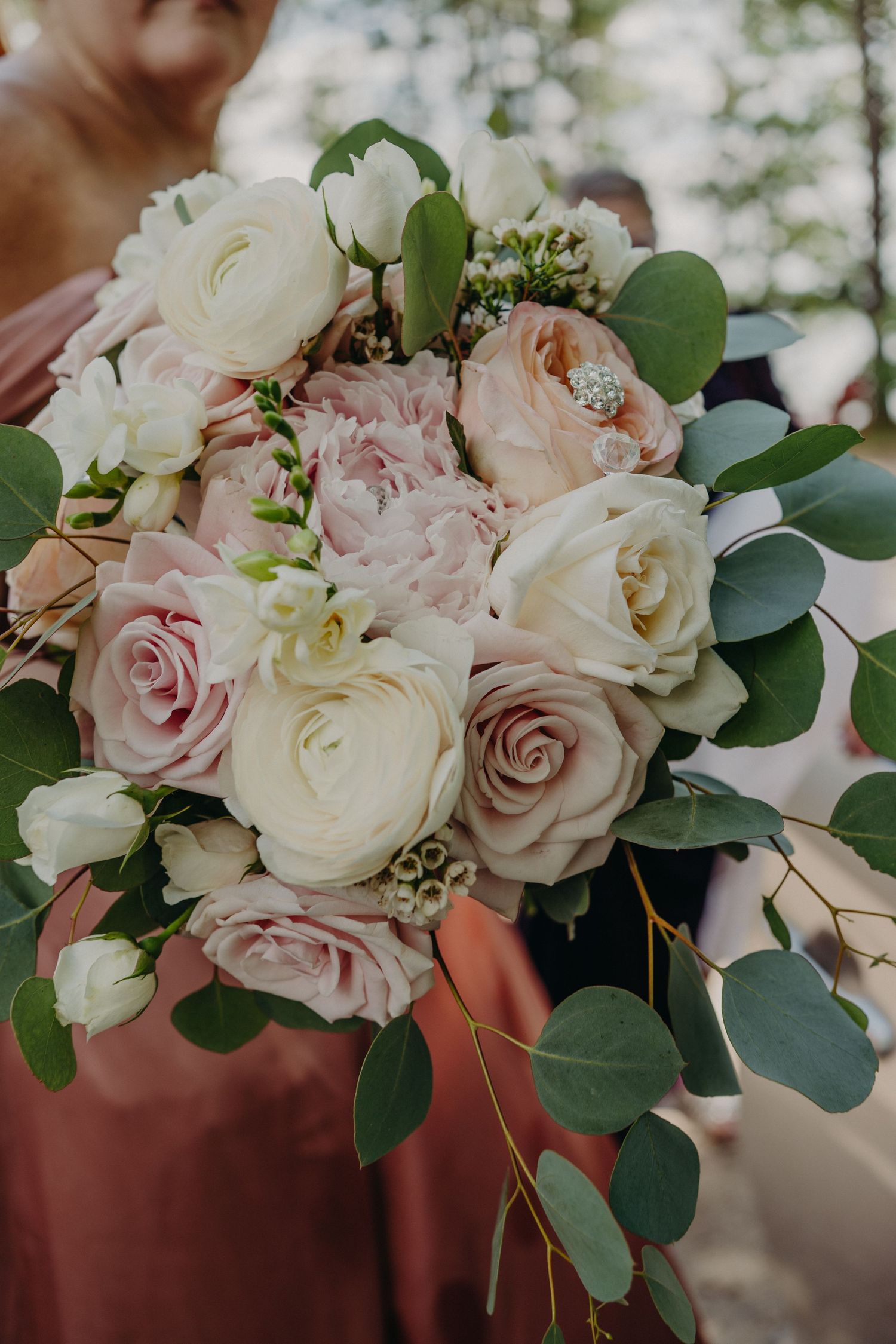 Elegant wedding bouquet featuring blush pink roses, white ranunculus, peonies, and eucalyptus held by a bridesmaid in dusty rose dress.