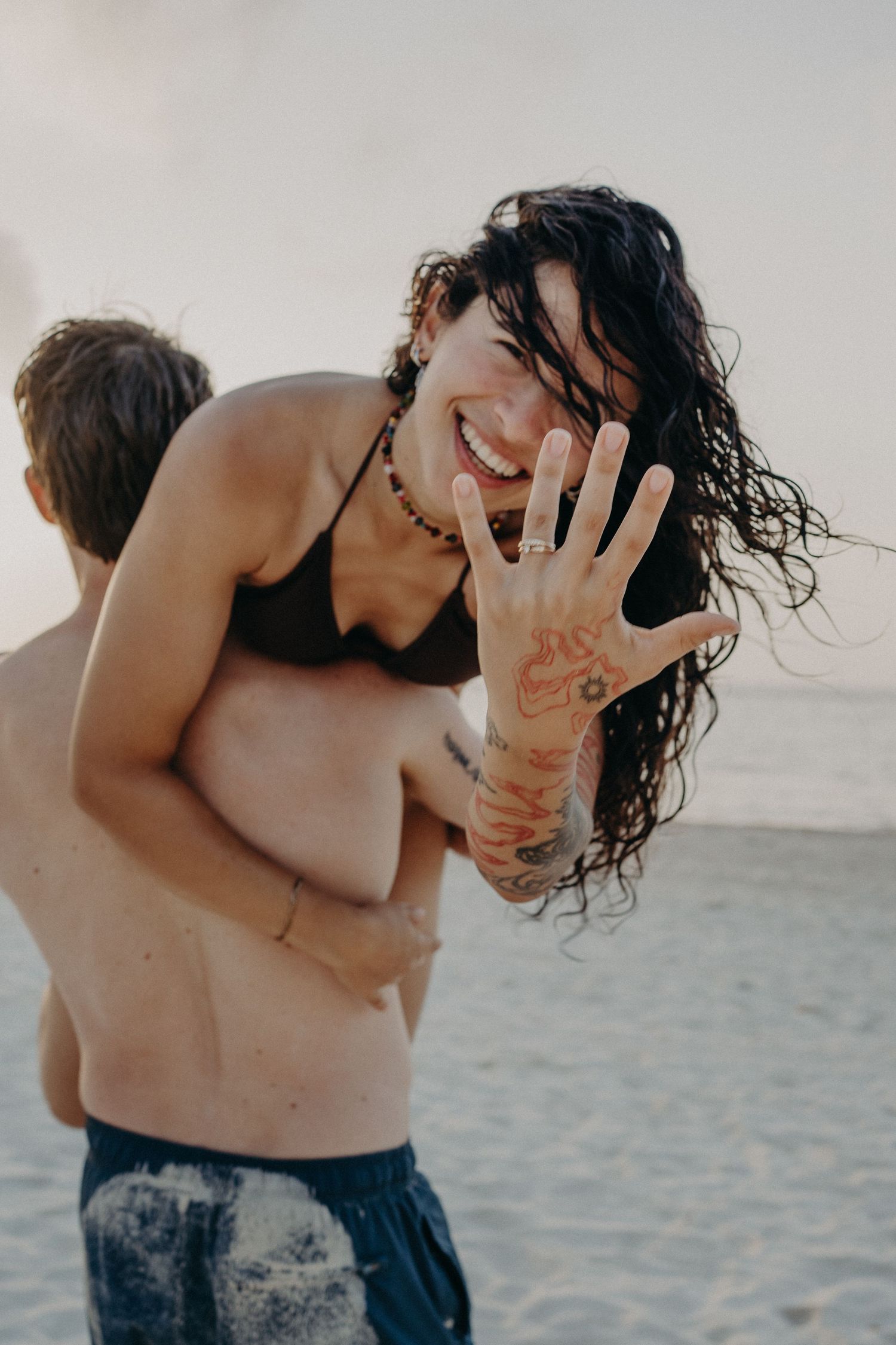 Happy woman in bikini showing engagement ring on beach, embraced by partner, with tattoos and wet hair.