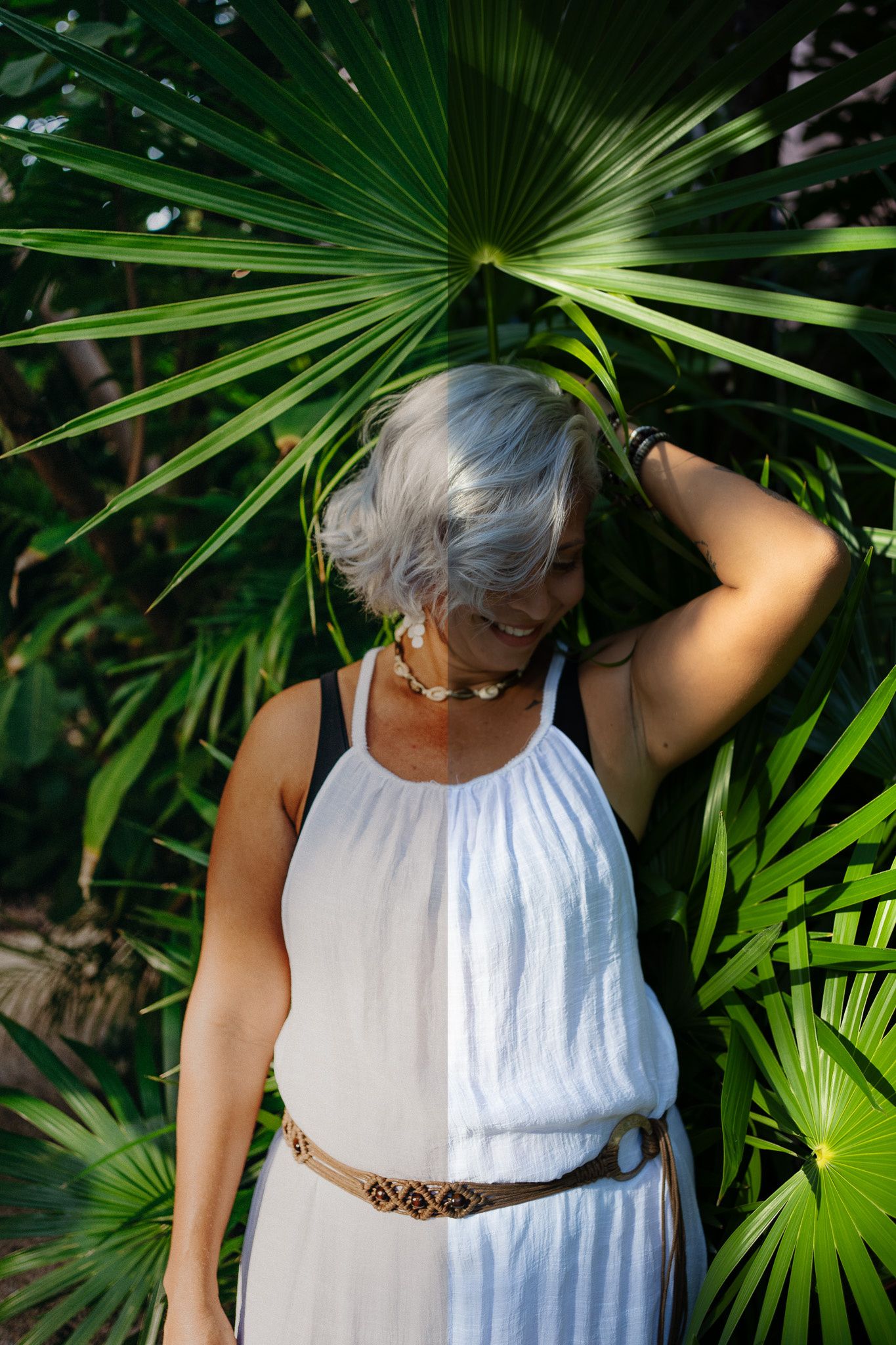 Smiling older woman with gray hair in white dress and belt posing among tropical palm leaves outdoors.