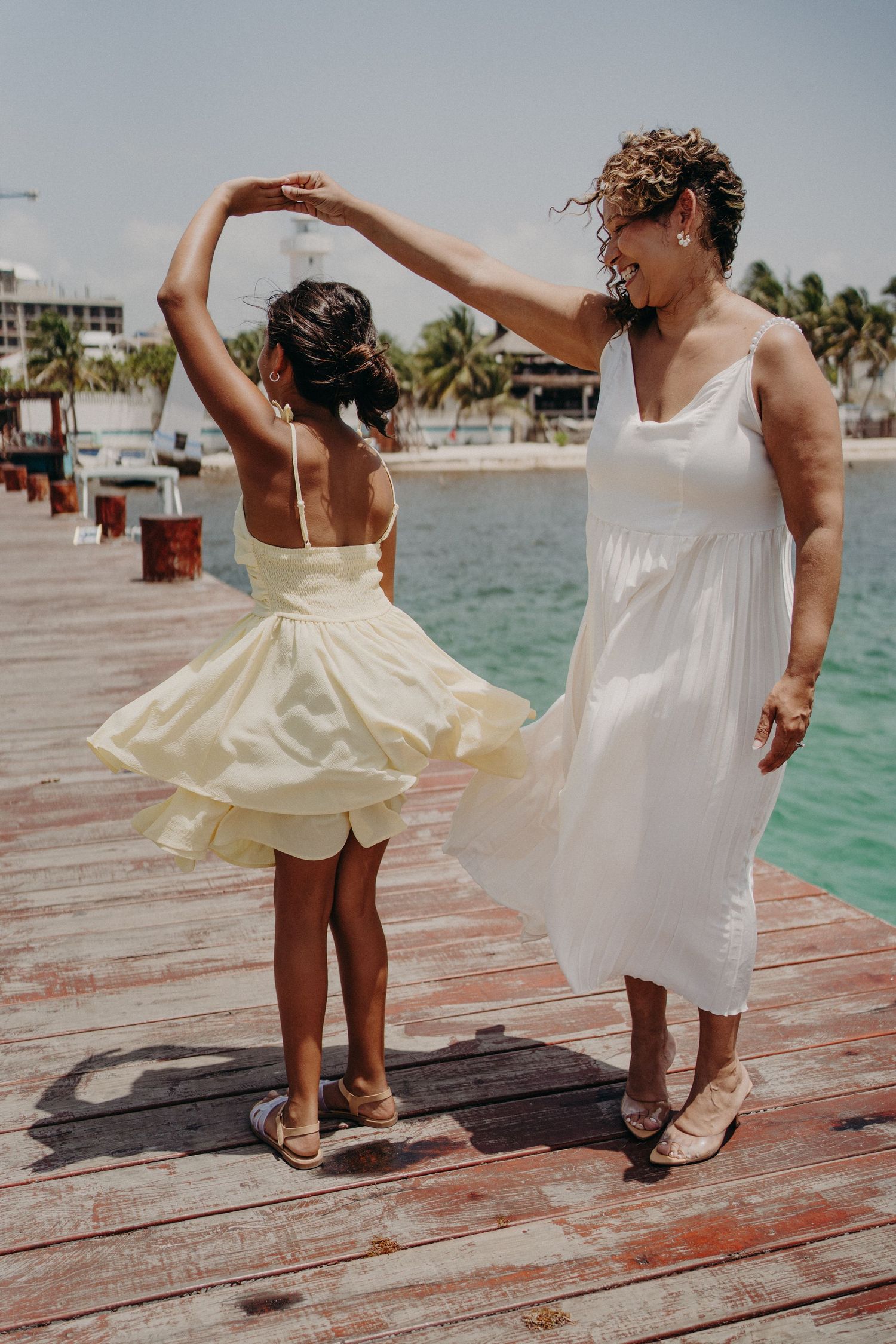 Woman in white dress twirling young girl in yellow dress on a wooden pier by turquoise water.
