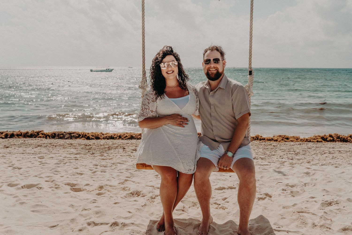 Couple smiling on a beach swing by the ocean, woman in white dress, man in casual shorts and shirt on a sunny day.