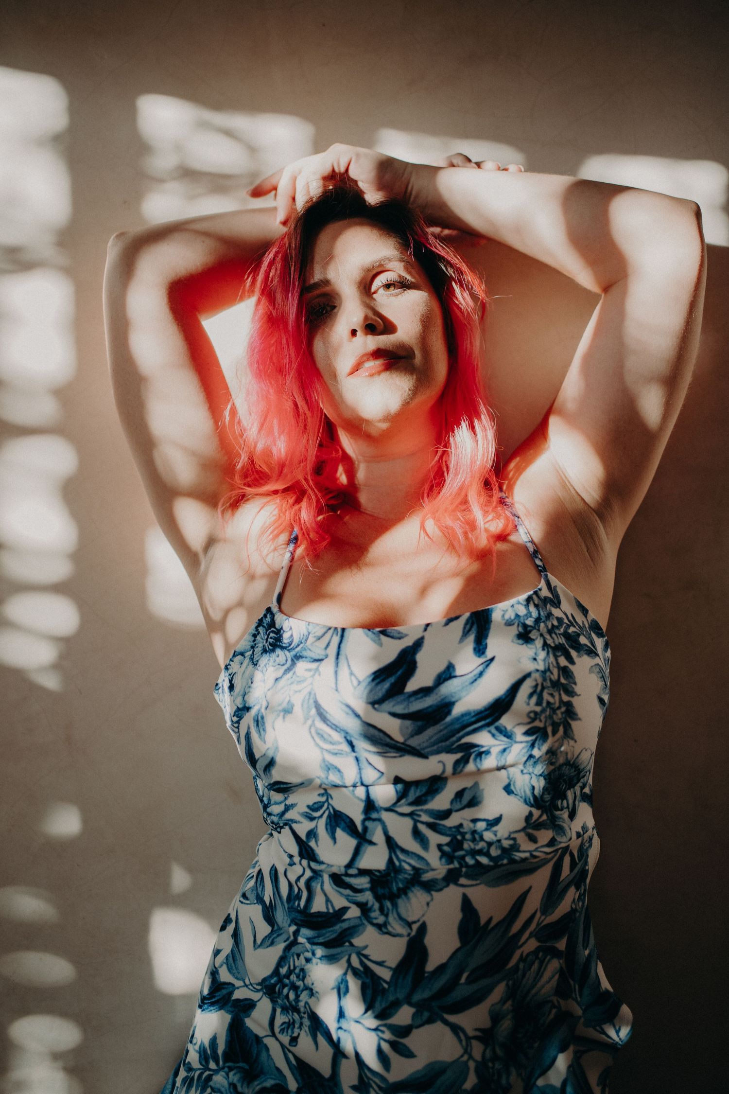 Woman with red hair in blue floral dress poses with arms raised against sunlit wall.