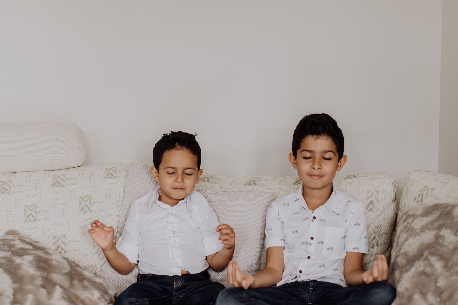 Two young boys in white shirts sit on a couch meditating with eyes closed in a calm, peaceful pose.