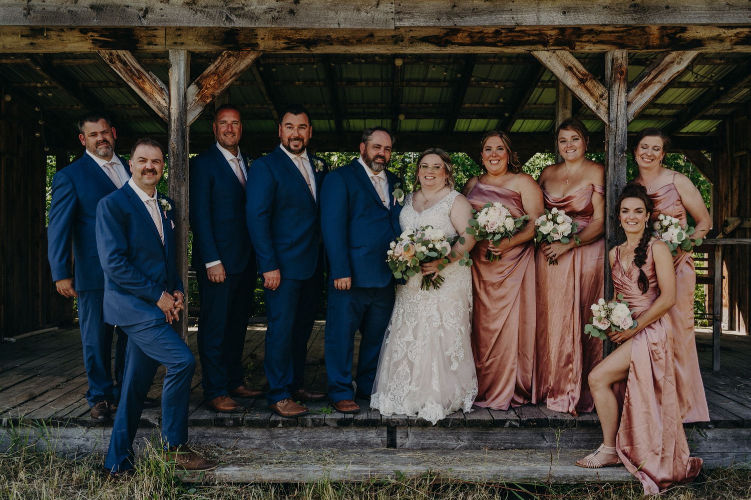 Wedding party posing under rustic wooden structure with groomsmen in navy and bridesmaids in rose.