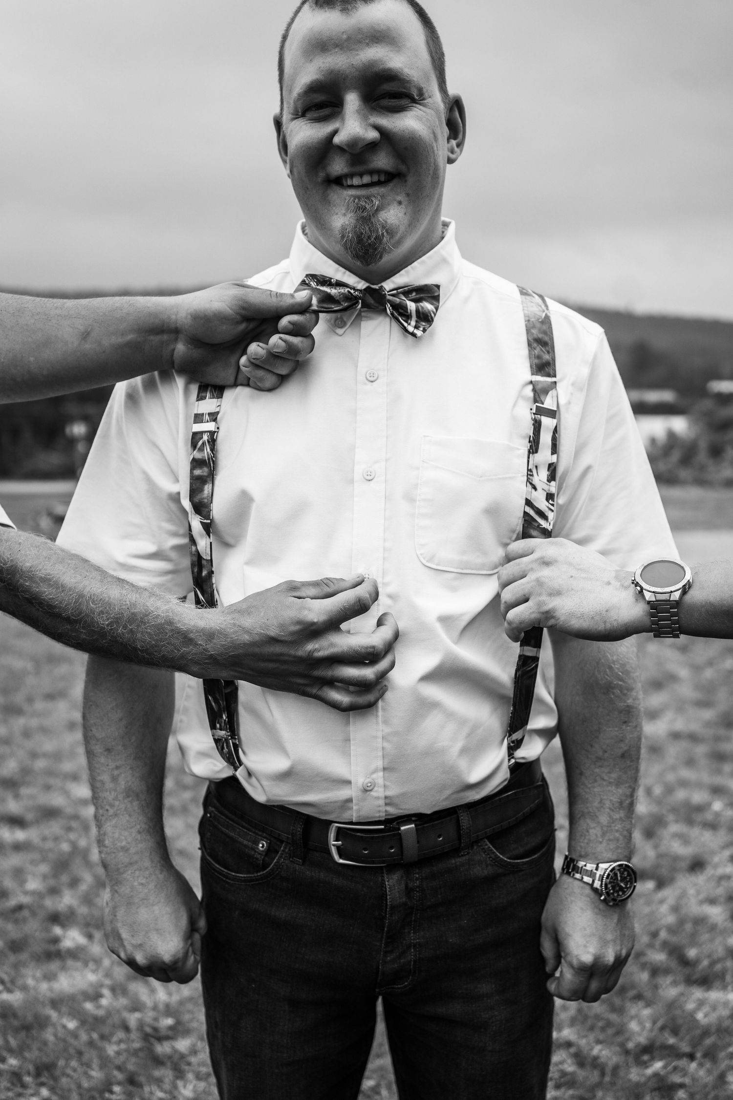 Groom smiling in white shirt with suspenders and bow tie being adjusted outdoors in black and white photo.