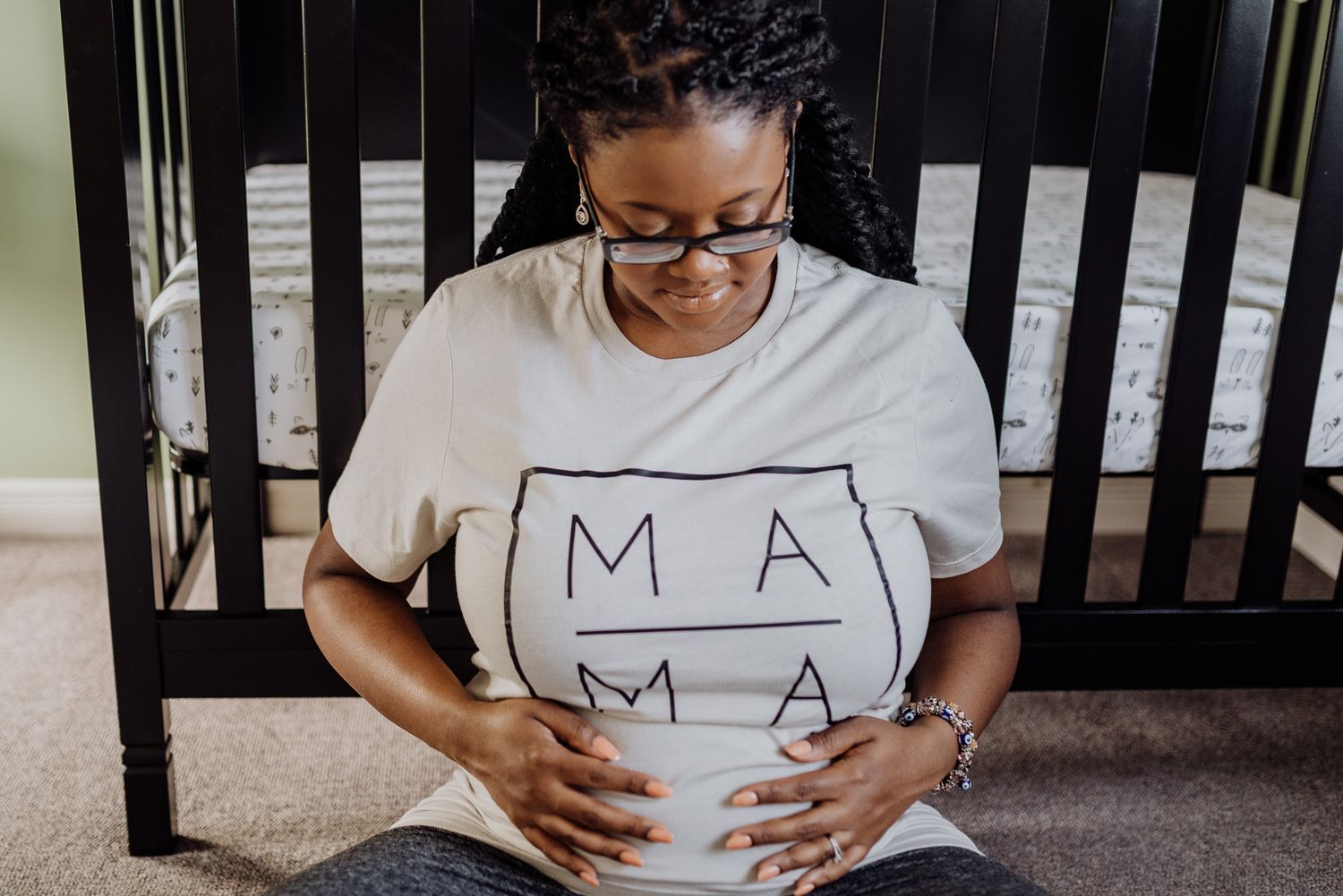 Pregnant woman in MAMA shirt sitting on floor by crib, cradling baby bump during maternity photo session.