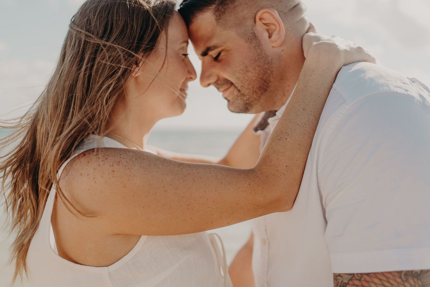 Romantic couple touching foreheads tenderly on a sunny beach, sharing an intimate moment together.