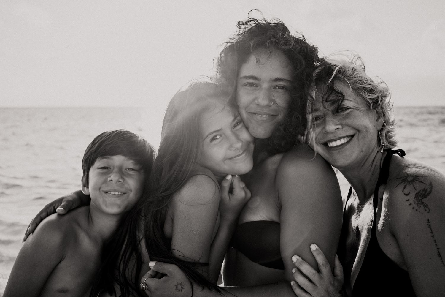 Black and white photo of four people hugging and smiling together on a beach near the ocean.