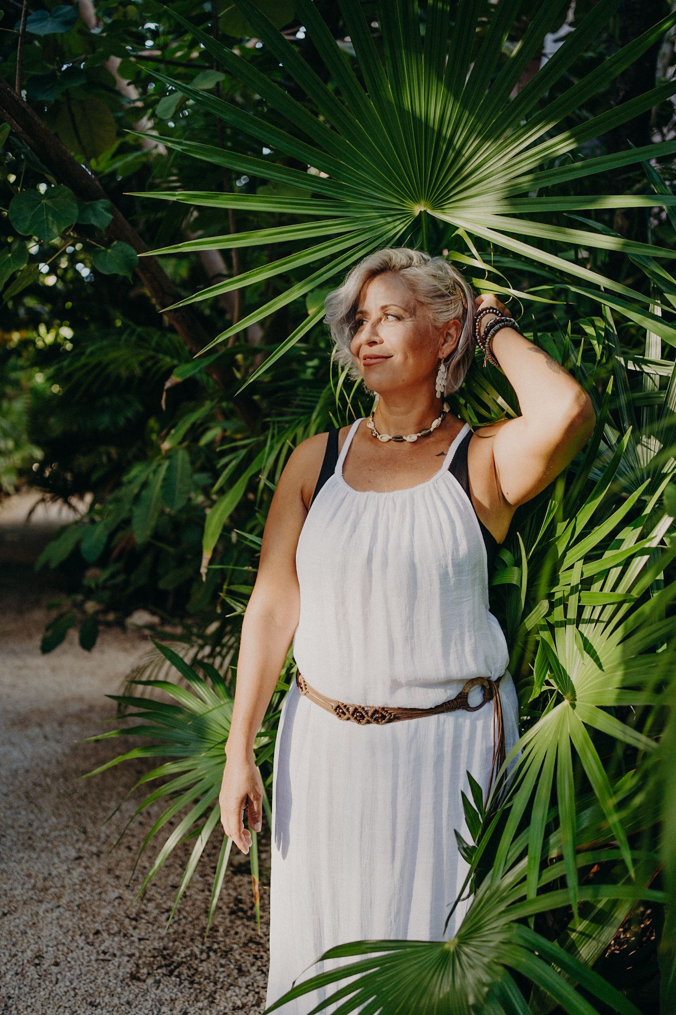 Woman in white dress and belt poses among lush tropical palm leaves in a serene outdoor setting.