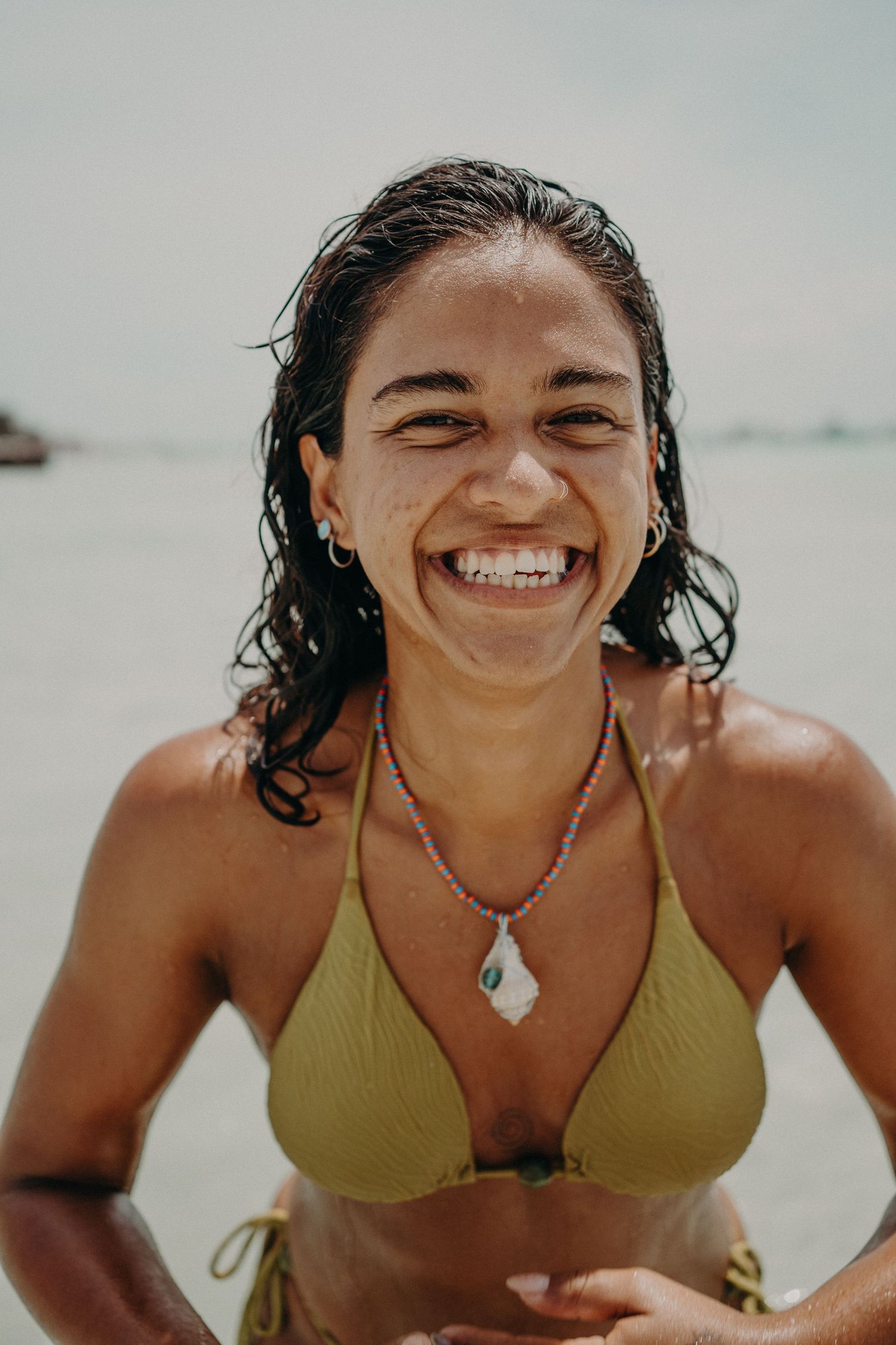 Happy young woman in yellow bikini smiling at beach, wearing beaded necklace with wet hair.