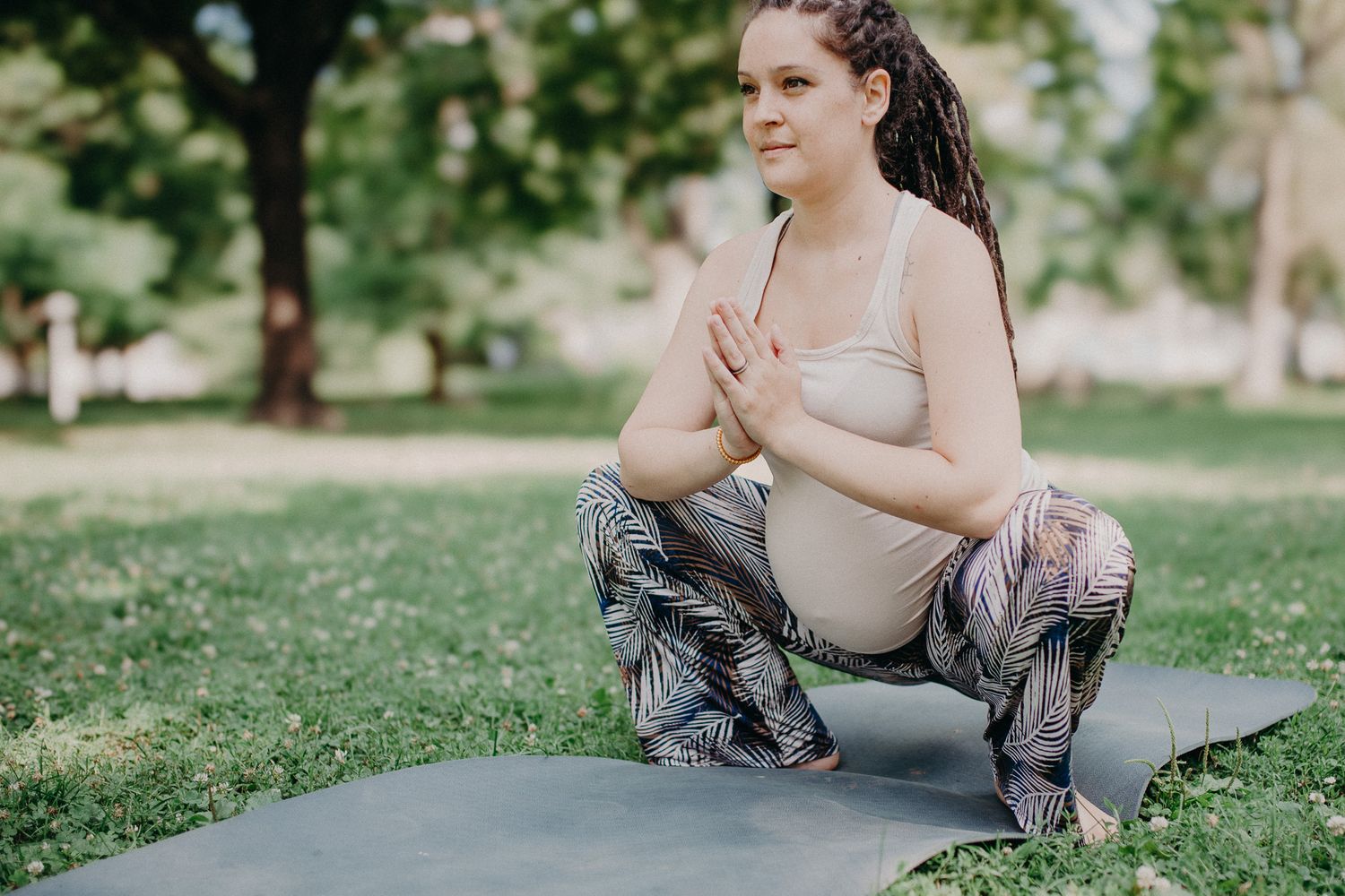 Pregnant woman doing yoga squat pose on mat in park, hands in prayer position, wearing patterned leggings.