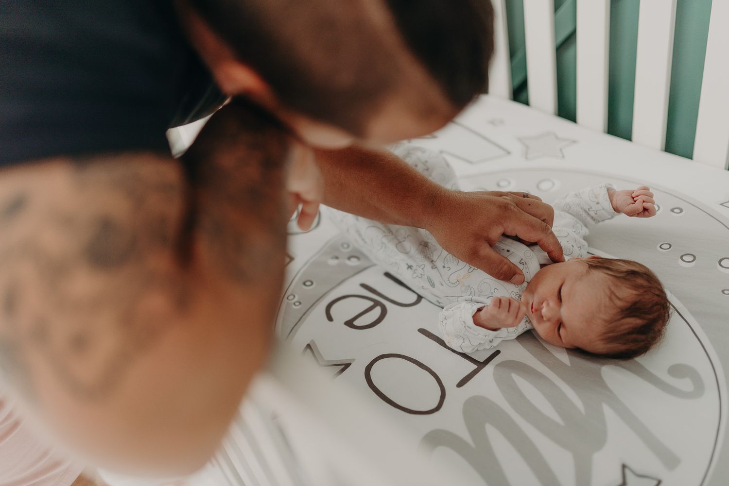 A tattooed parent leans over a crib to tend to their sleeping newborn baby.