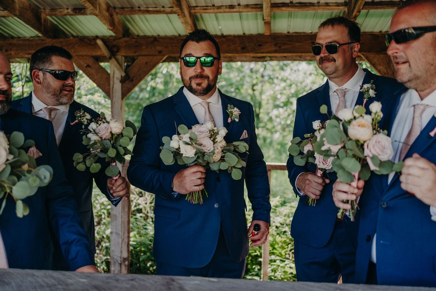 Groomsmen in navy blue suits holding white floral bouquets with eucalyptus under a wooden pavilion.