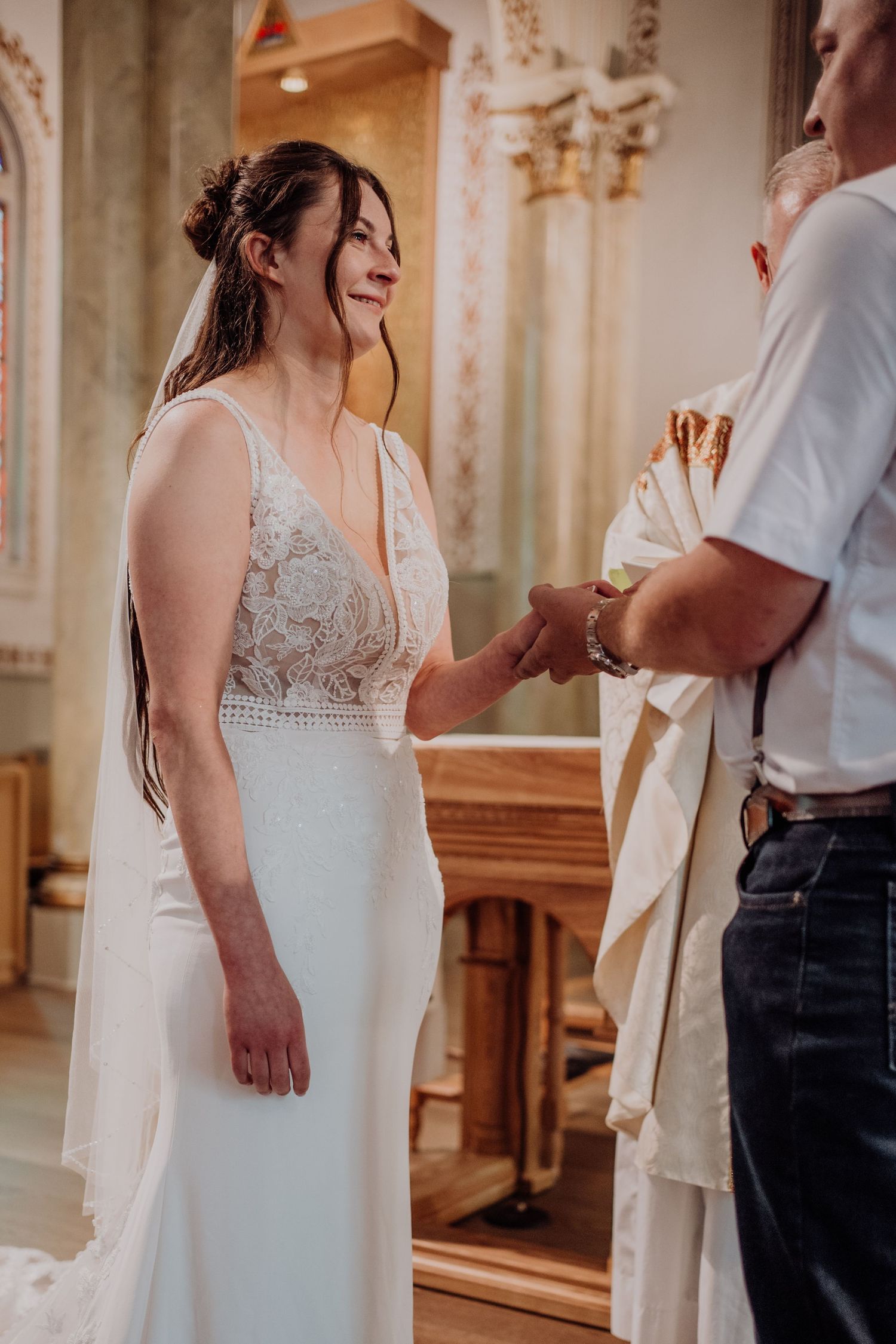 Bride in lace wedding dress smiling while holding grooms hands during church ceremony at altar.