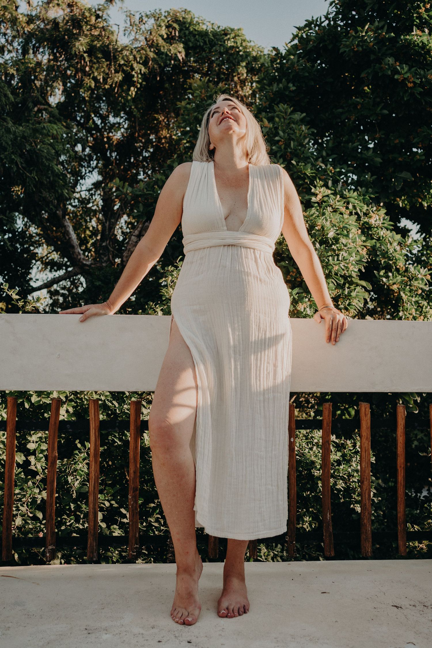 Blonde woman in elegant white ribbed midi dress with deep V-neck stands on sunlit balcony surrounded by trees.