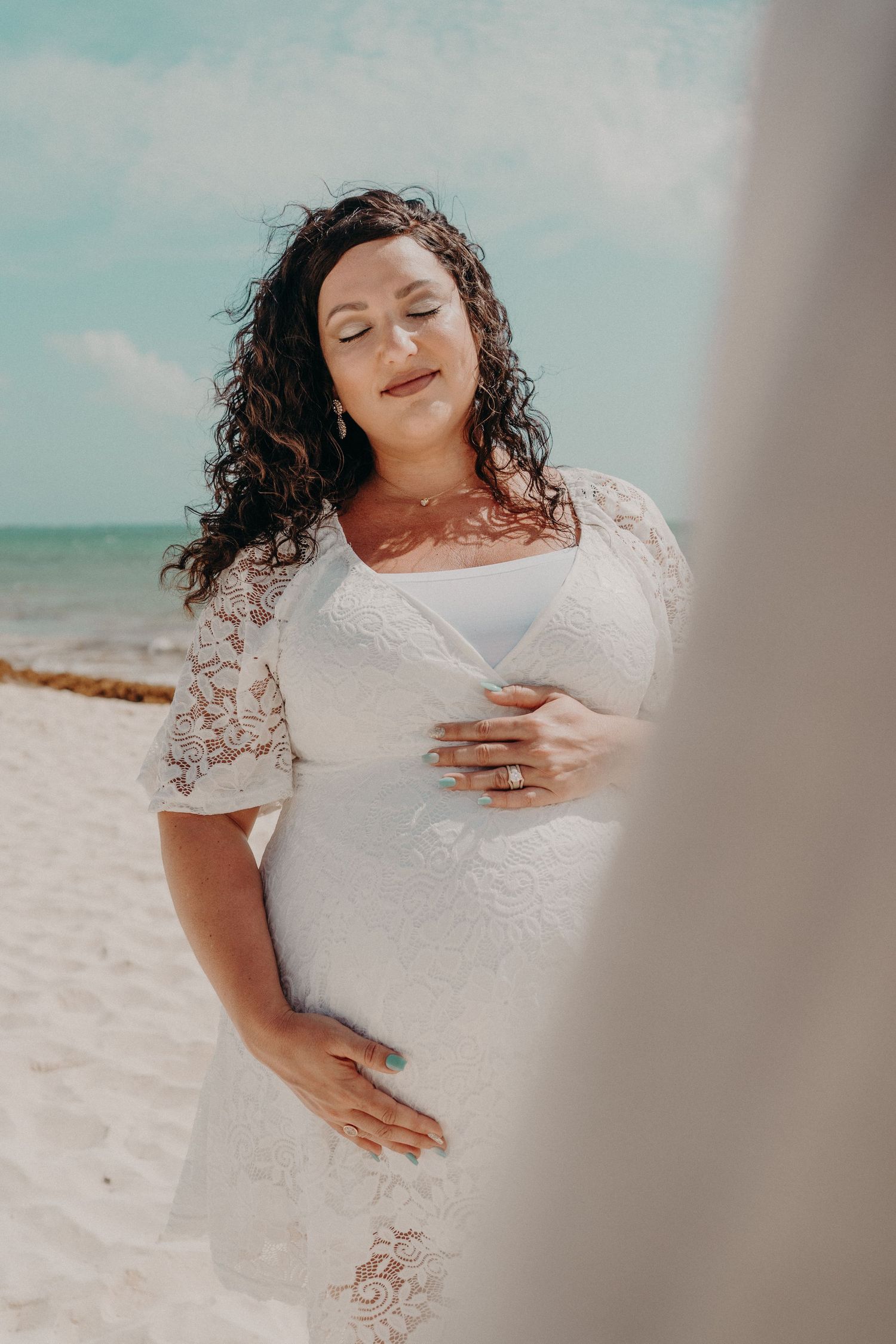 Pregnant woman in white lace dress holding belly on sandy beach, eyes closed, serene expression, ocean in background.