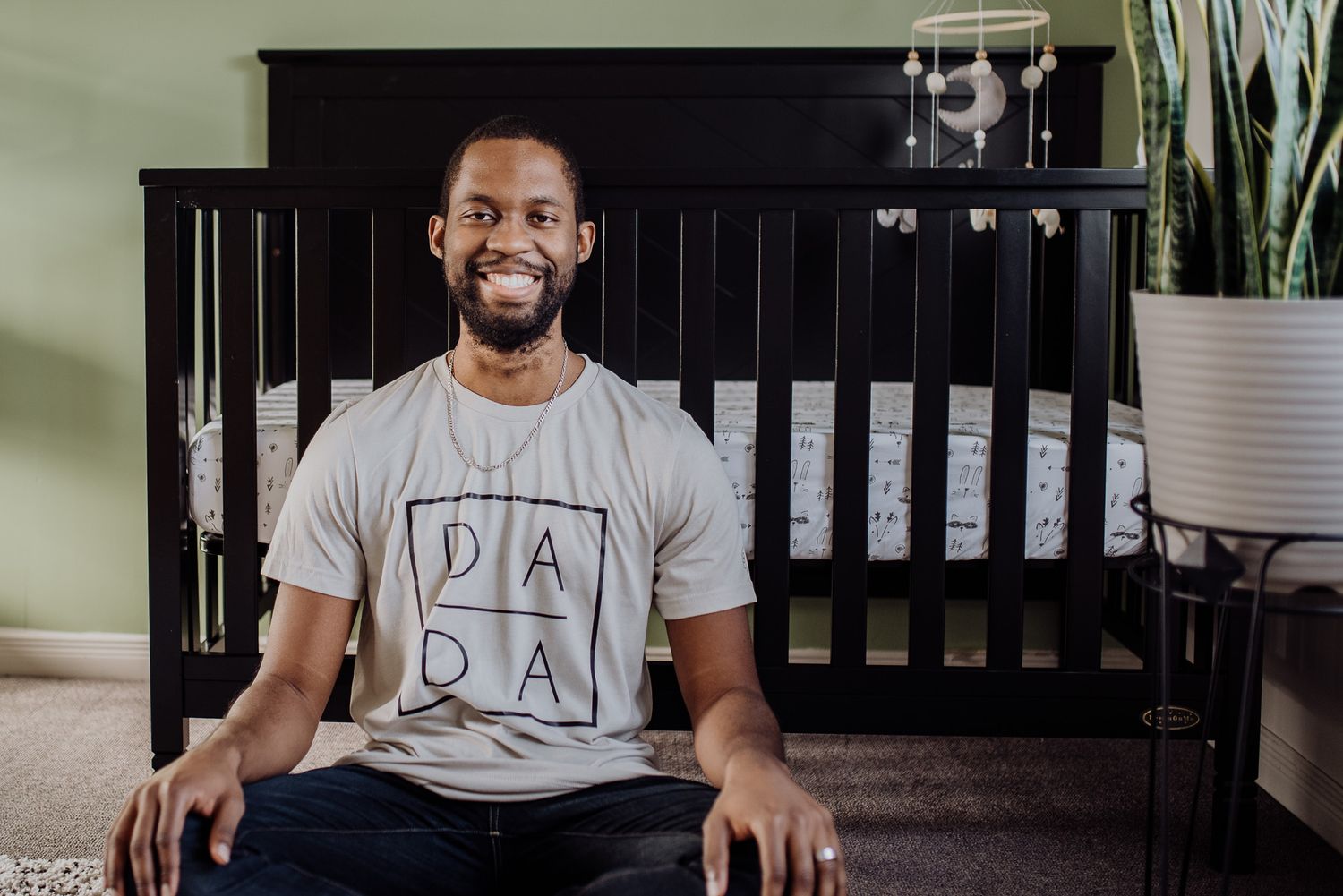 Smiling man wearing a DADA t-shirt sits cross-legged in front of a dark wood baby crib in a nursery.