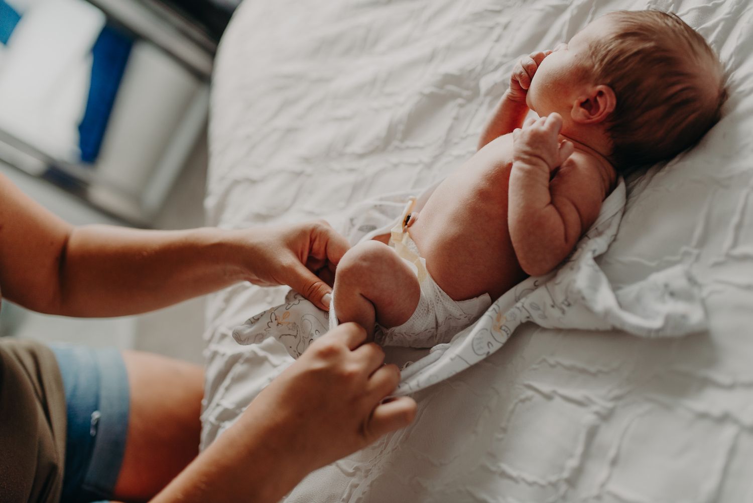 Parent changing diaper of newborn baby lying on white bed, tender care moment captured from above.
