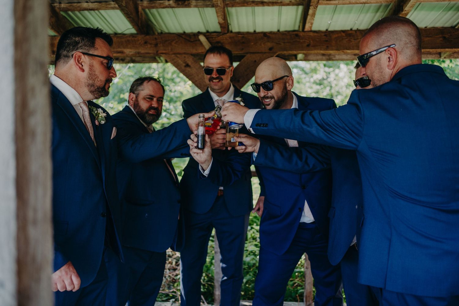 Groomsmen in navy suits toasting with drinks under a wooden outdoor pavilion at a wedding.