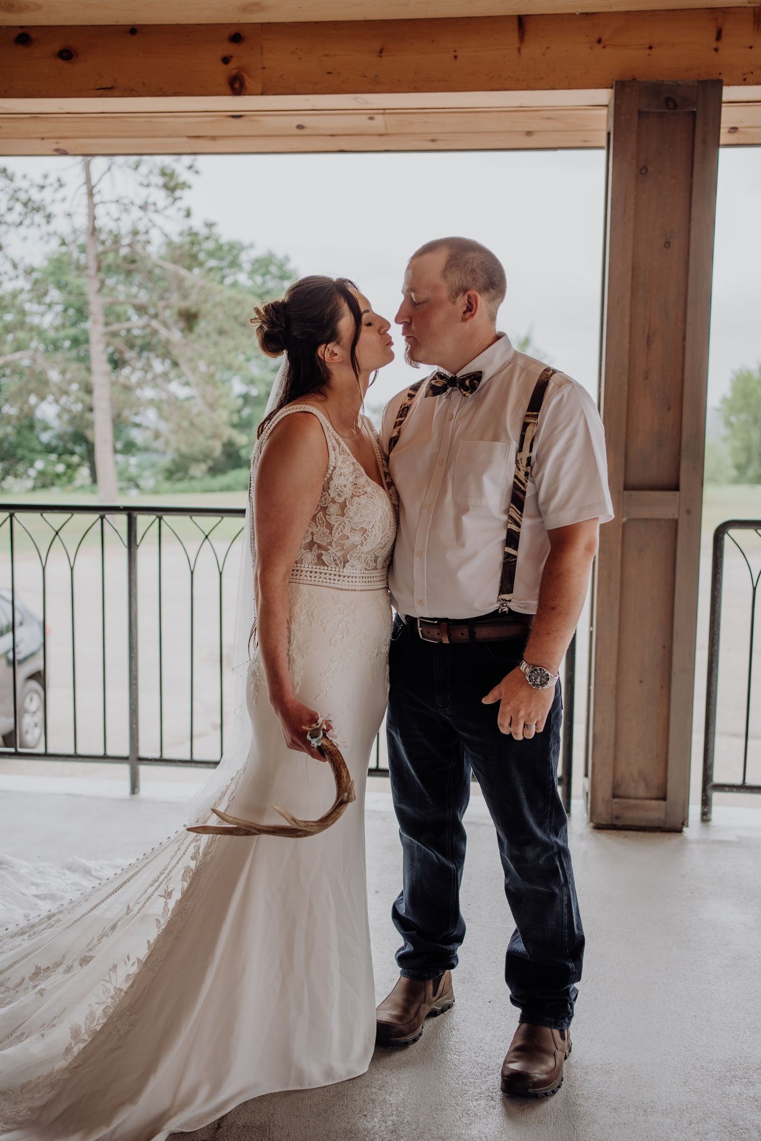 Bride and groom sharing a kiss on covered porch, groom wearing suspenders and bow tie.