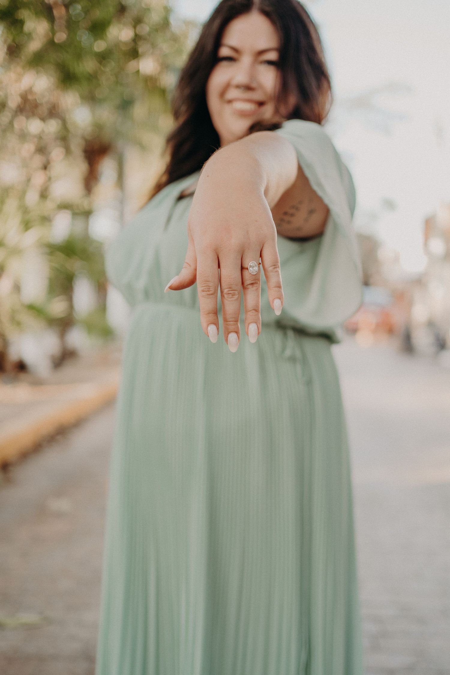 Smiling woman in mint green pleated maxi dress extending hand showing diamond engagement ring outdoors.
