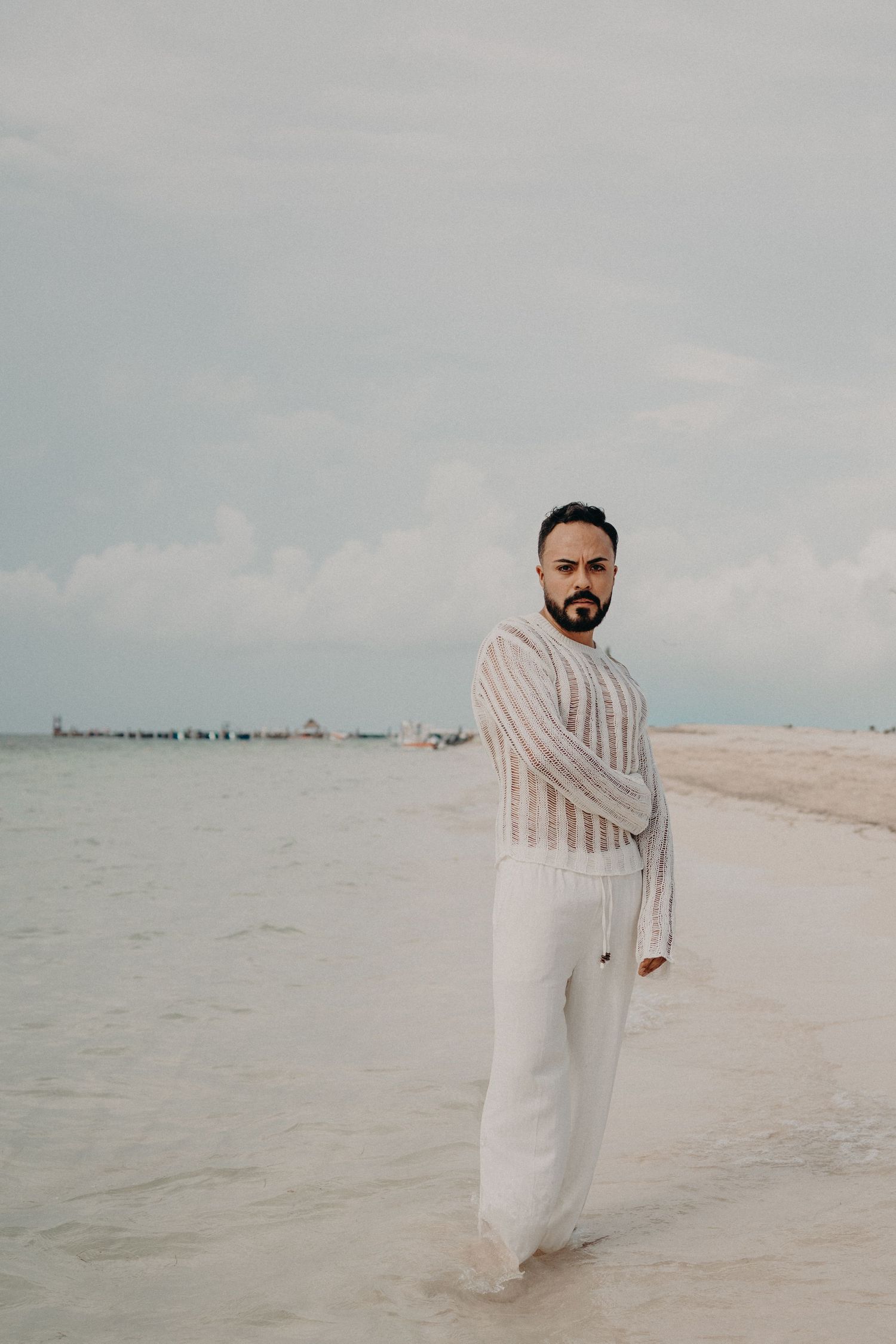 A bearded man in white linen pants and sheer top stands on a tropical beach with calm turquoise waters behind him.