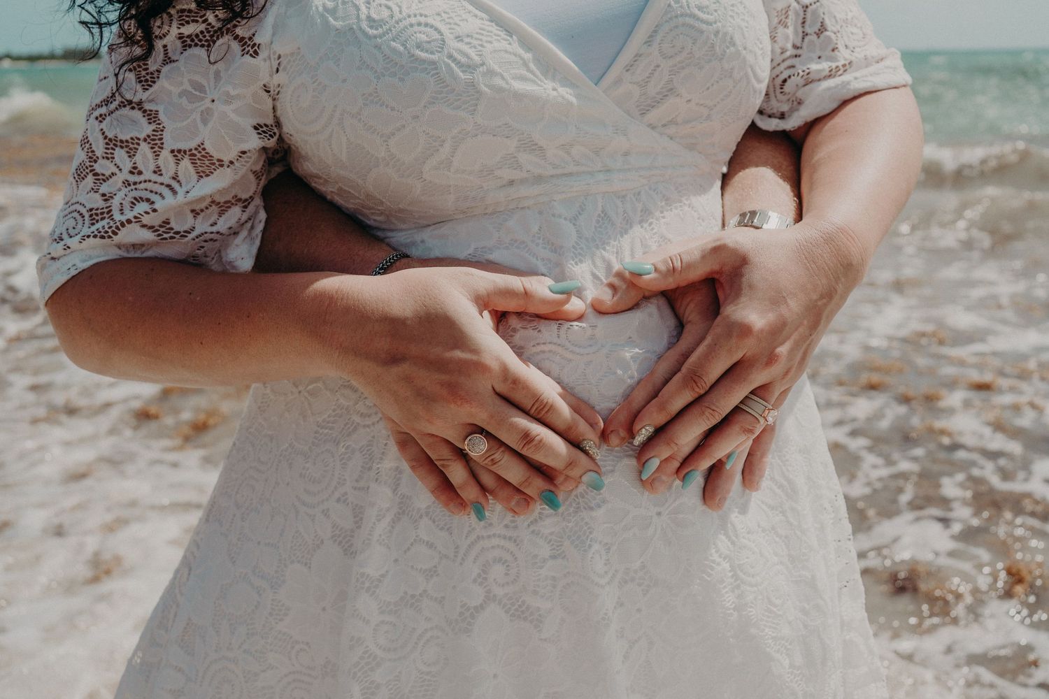 Pregnant bride in lace wedding dress on beach with partner forming heart shape with hands on baby bump.