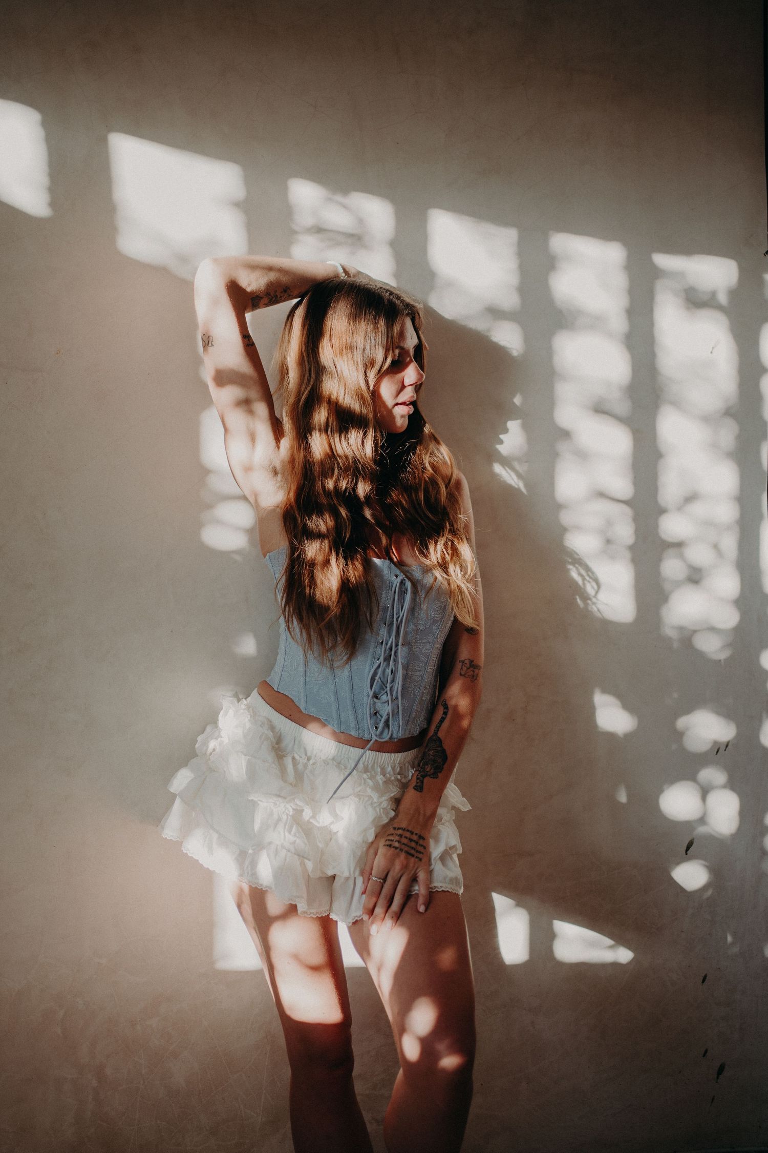 Young woman with long wavy hair poses in blue corset top and white ruffled mini skirt against sunlit wall.