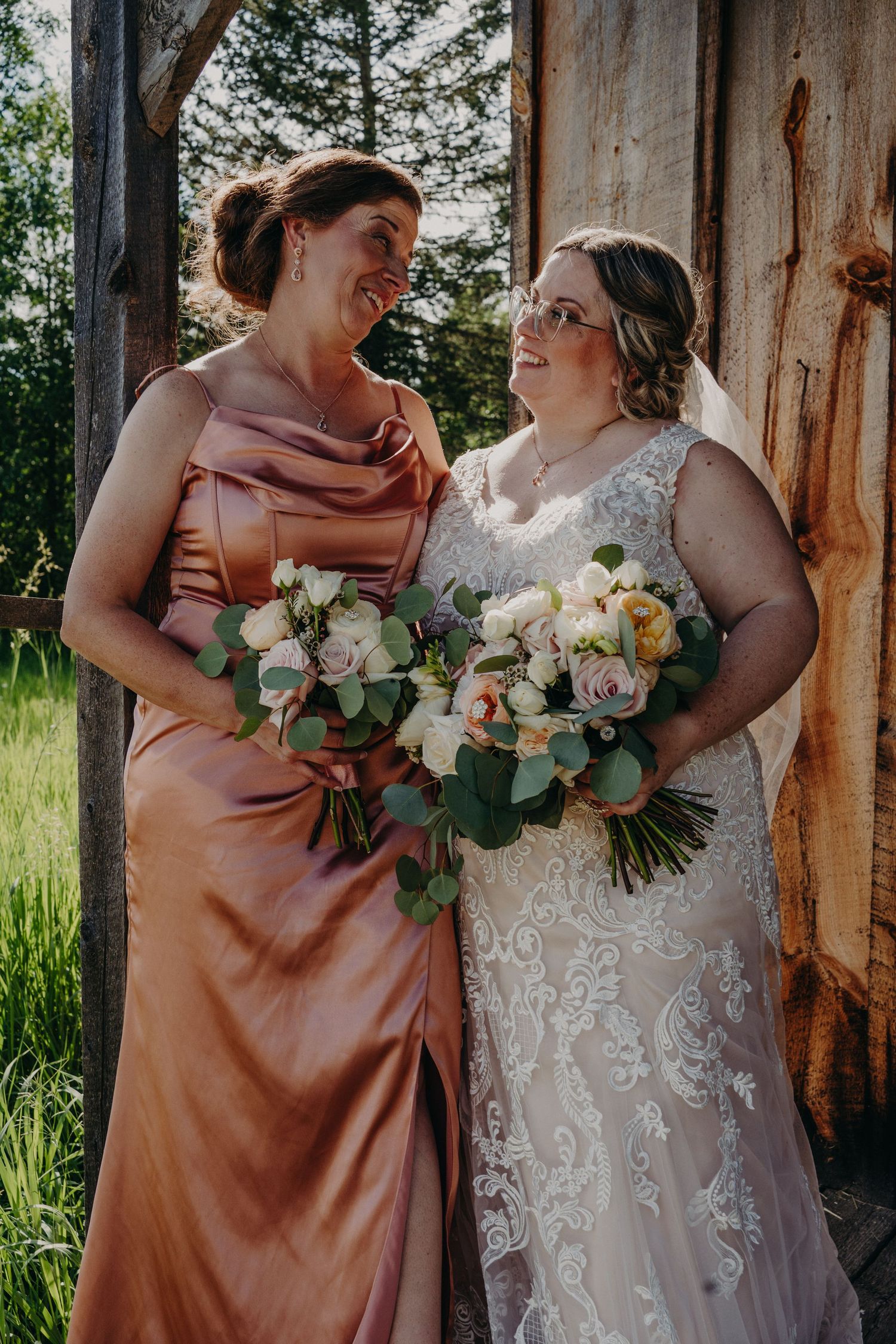 Bride in lace gown and bridesmaid in dusty rose satin dress sharing a joyful moment outdoors.