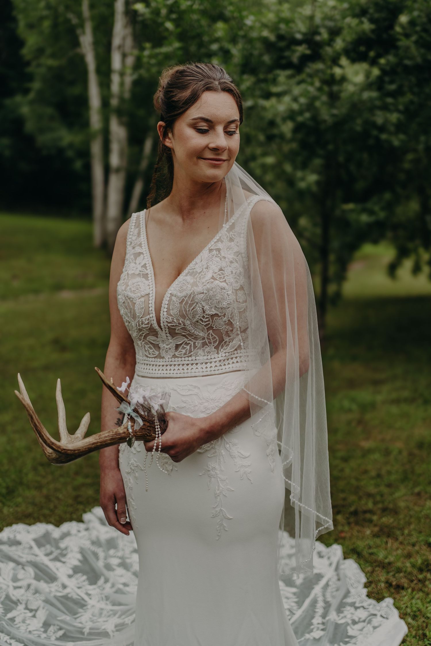 Beautiful bride in lace wedding gown holding deer antlers with veil in lush green outdoor setting.