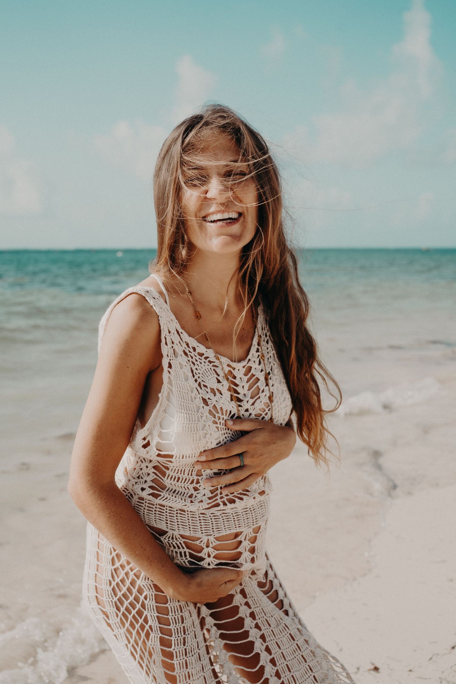 Smiling woman with long windswept hair wearing a white crochet beach cover-up on a sandy tropical beach with turquoise ocean.