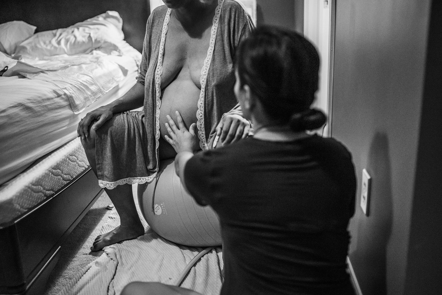 Black and white photo of a pregnant woman sitting on a bed being comforted by a support person during labor.