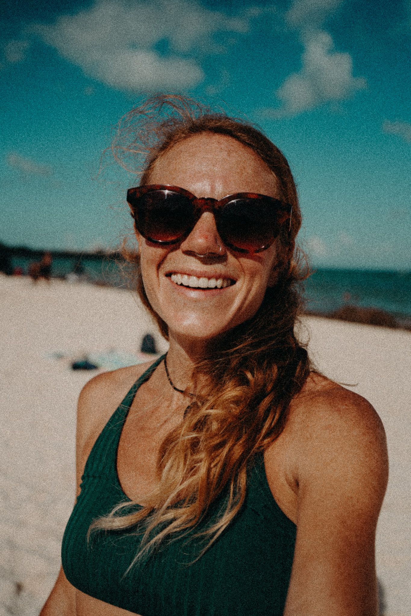 Smiling woman with sunglasses and green bikini top on sunny beach with blue sky.