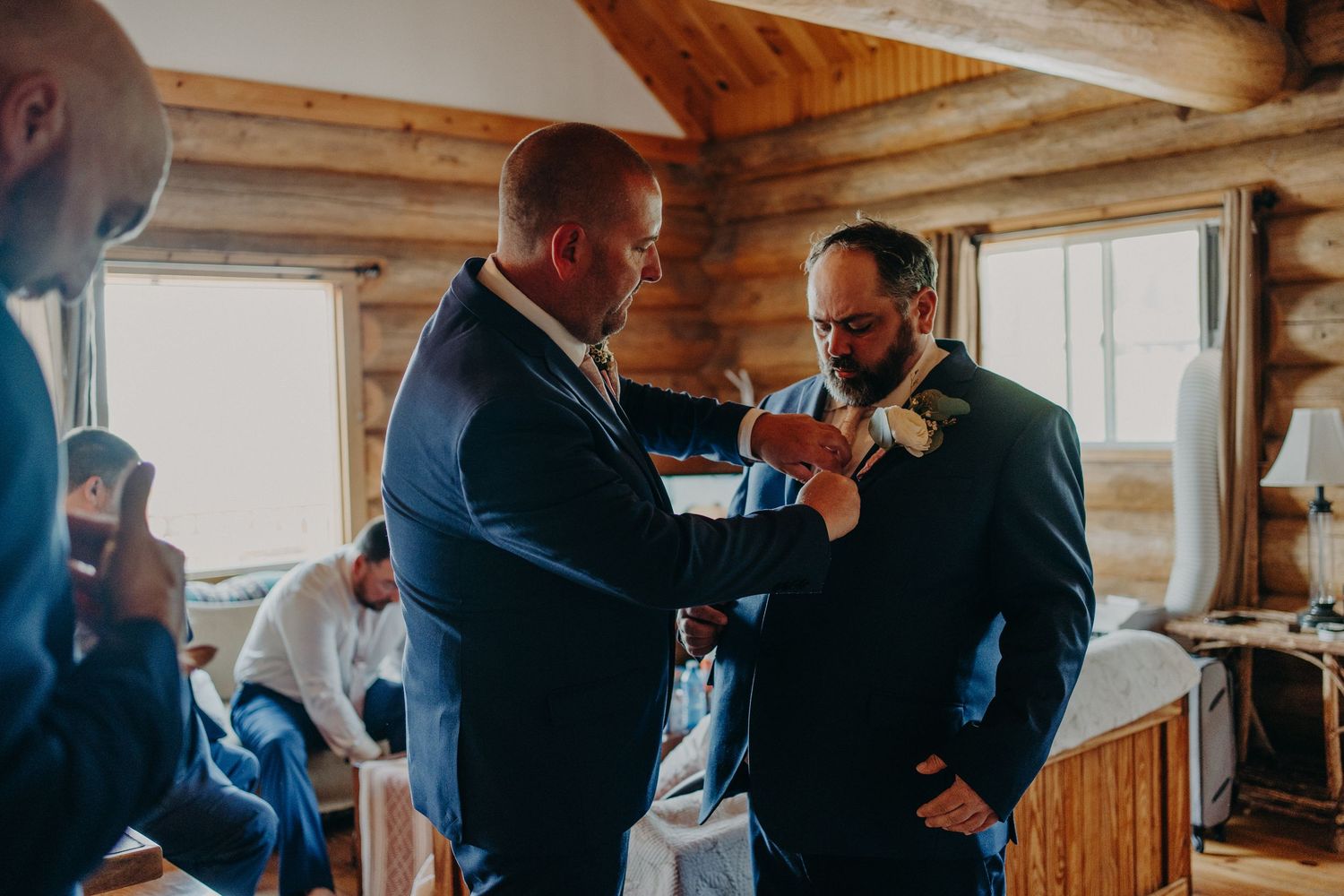 Groomsmen in navy suits helping groom with boutonniere in rustic log cabin wedding venue.
