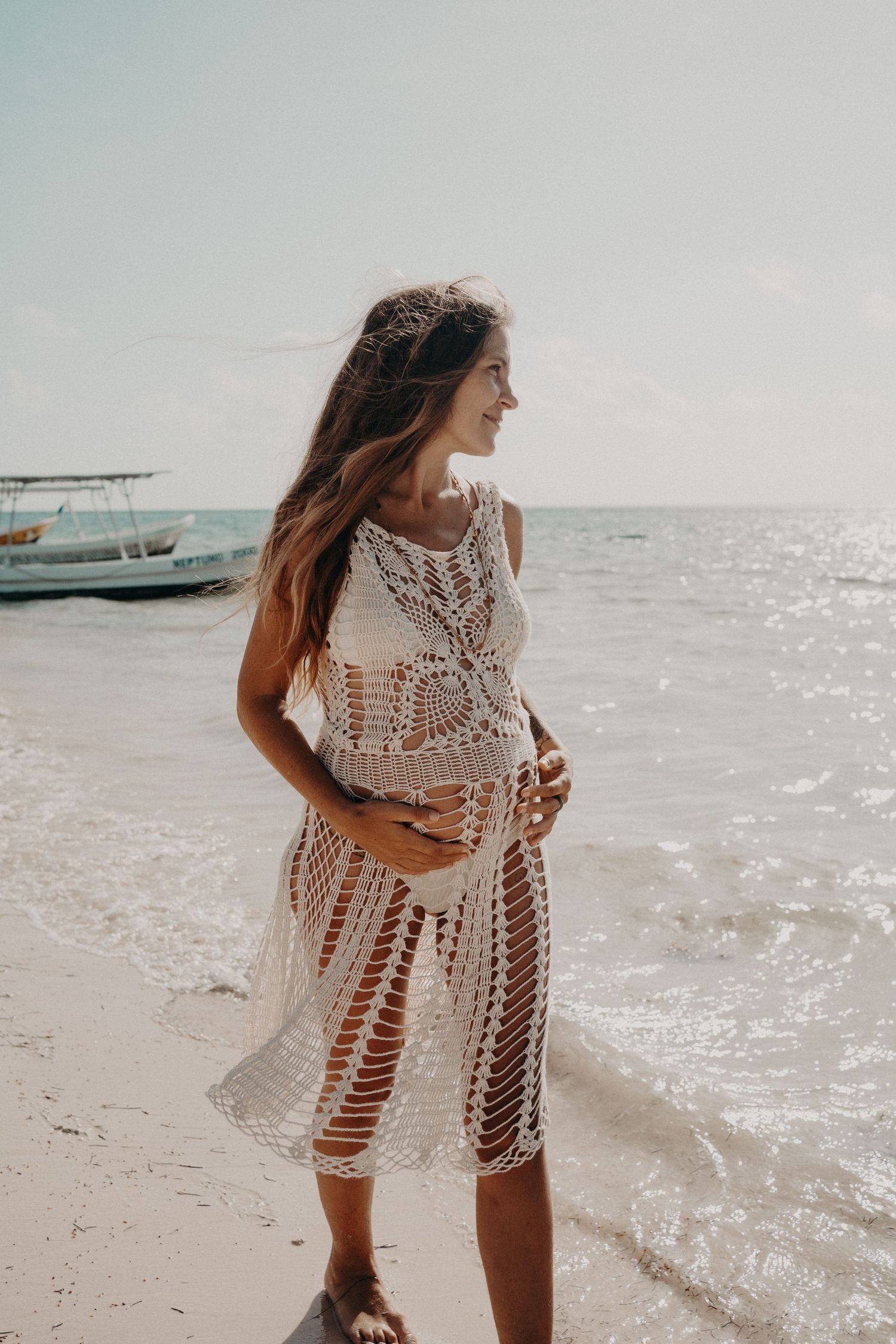 Woman in a white crochet beach dress standing on a sunny shoreline, hands on her belly, with a boat and calm ocean behind her.