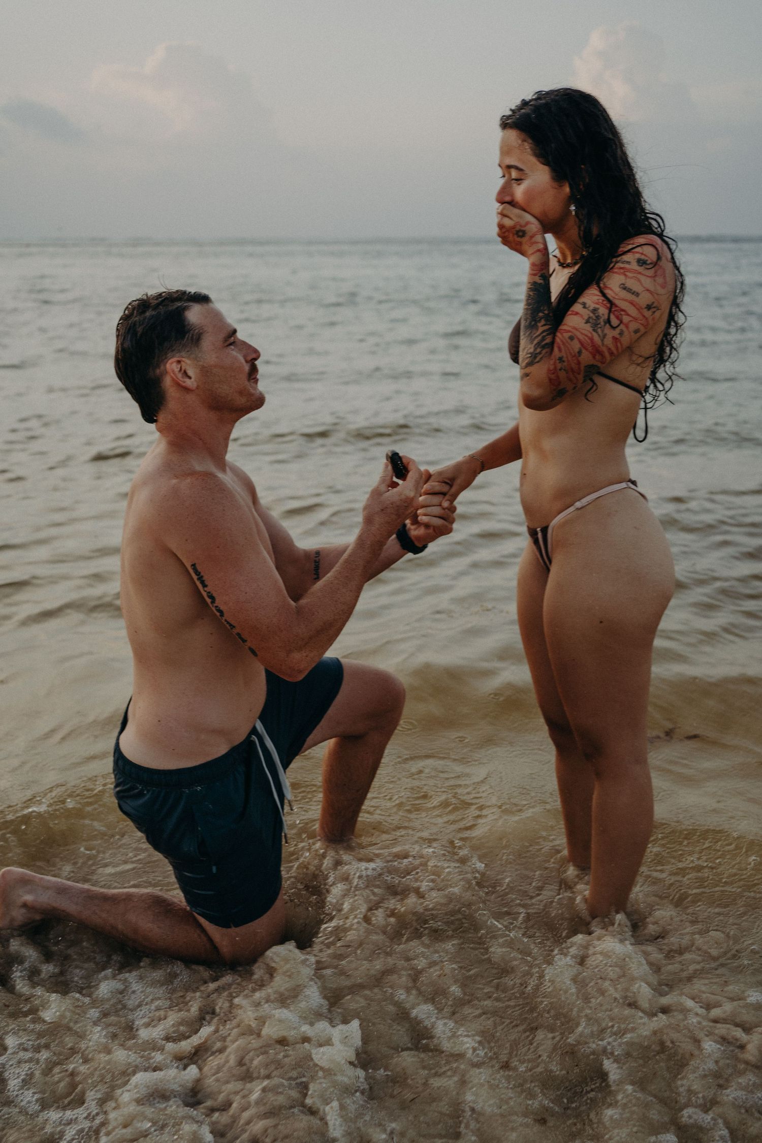 Man proposes on one knee to woman in bikini at ocean shore during sunset beach marriage proposal.