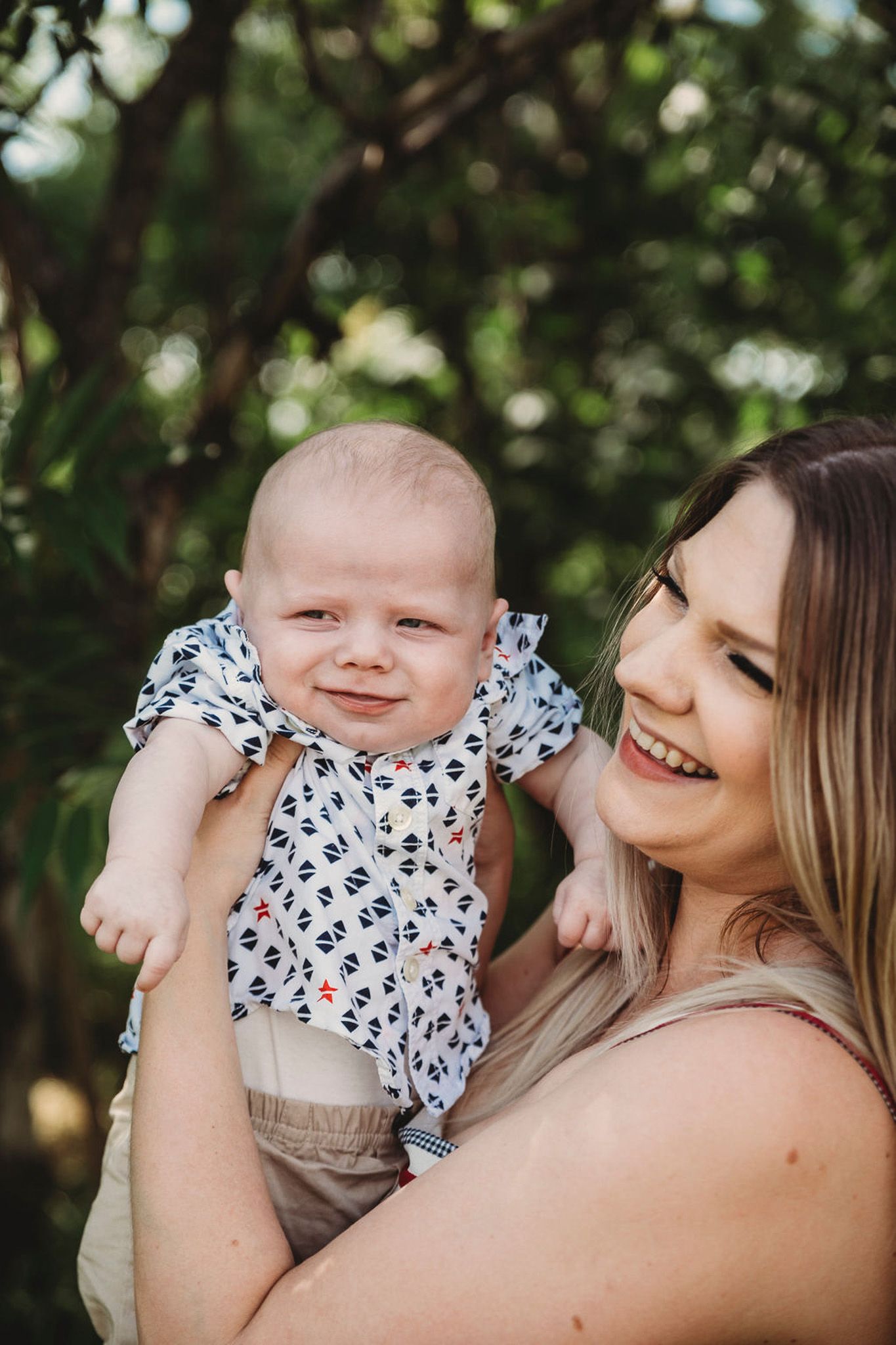 A smiling mother holds her happy baby outdoors surrounded by lush green trees.