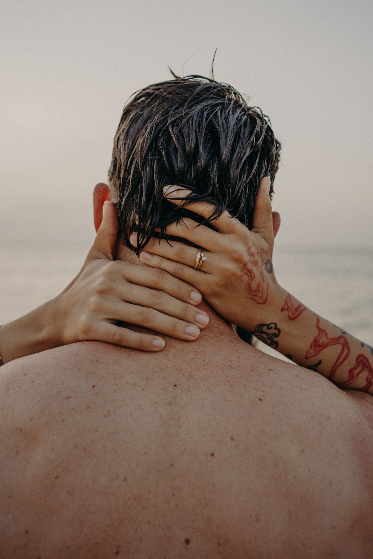 Two arms with red tattoos and a ring embrace the neck of a shirtless man with wet dark hair, seen from behind at a hazy beach.