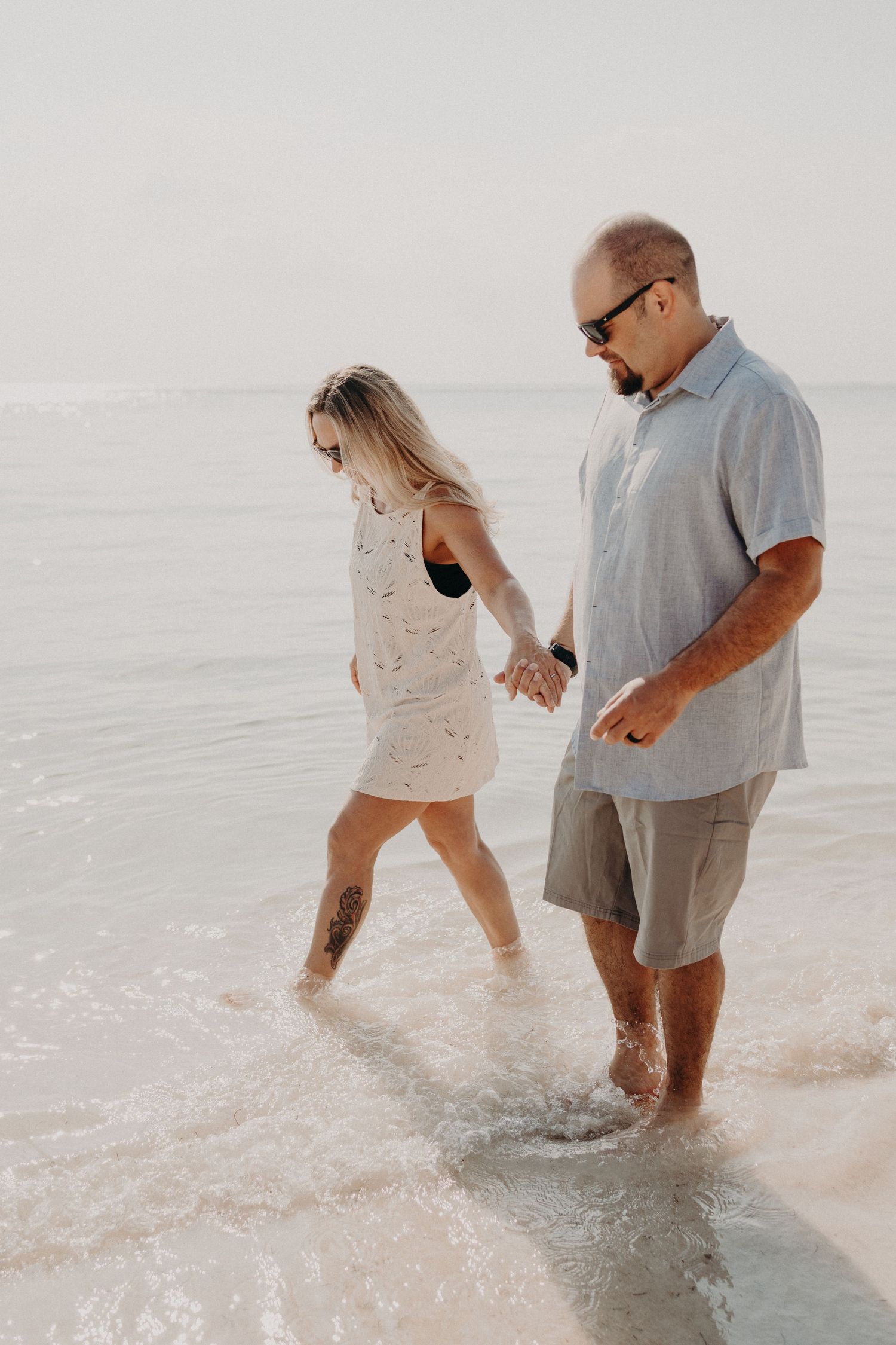 A couple holds hands while wading through shallow ocean waves on a sandy beach, enjoying a romantic seaside stroll.