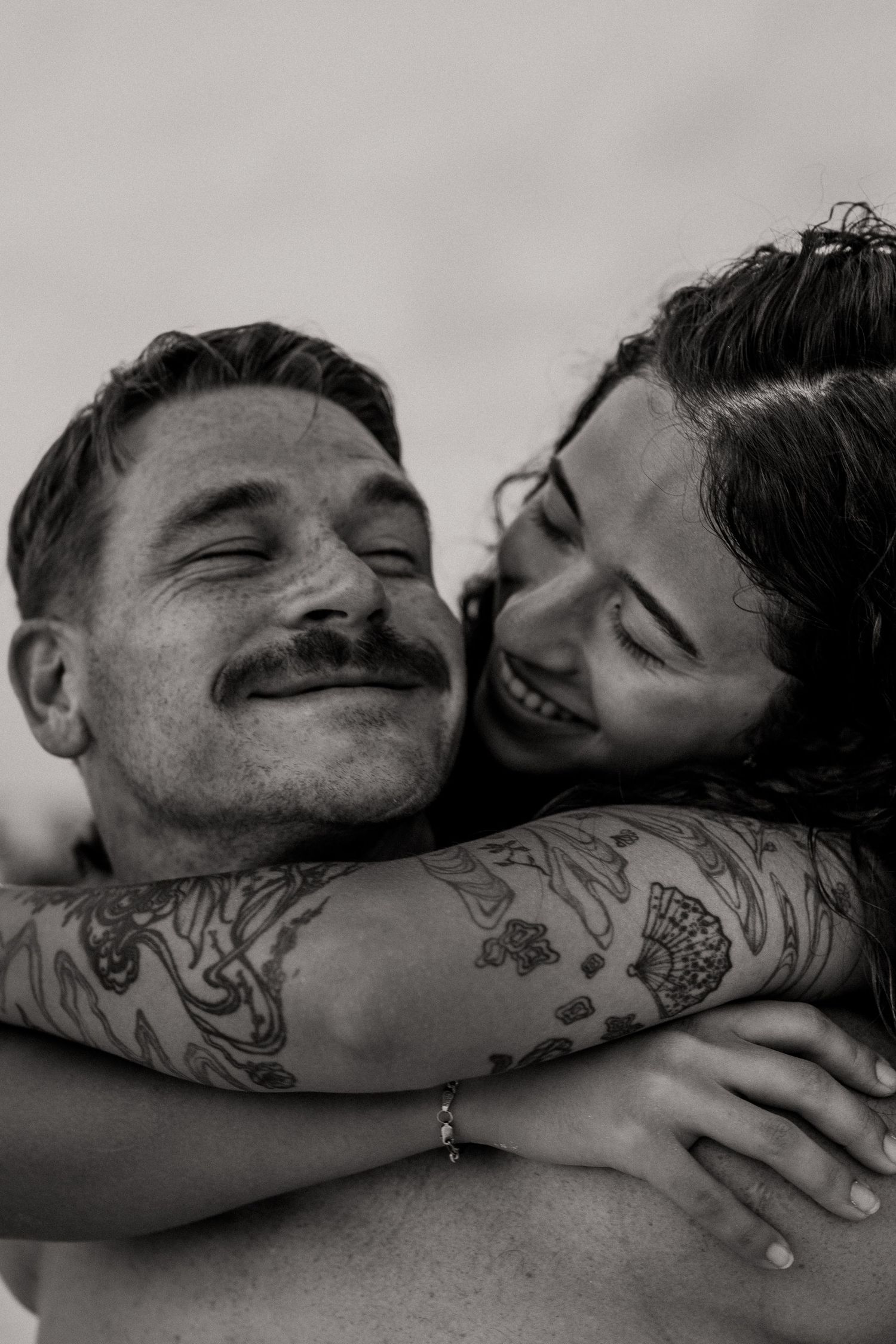 Black and white photo of a smiling couple embracing, man with mustache and tattoos, woman hugging him from behind.