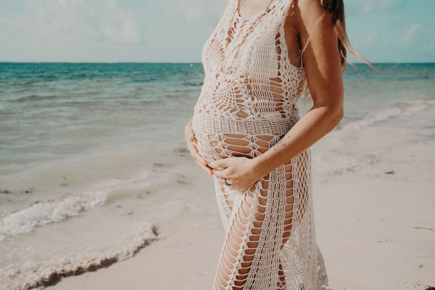 Pregnant woman in a white crochet cover-up cradling her baby bump on a sandy tropical beach beside turquoise ocean waves.