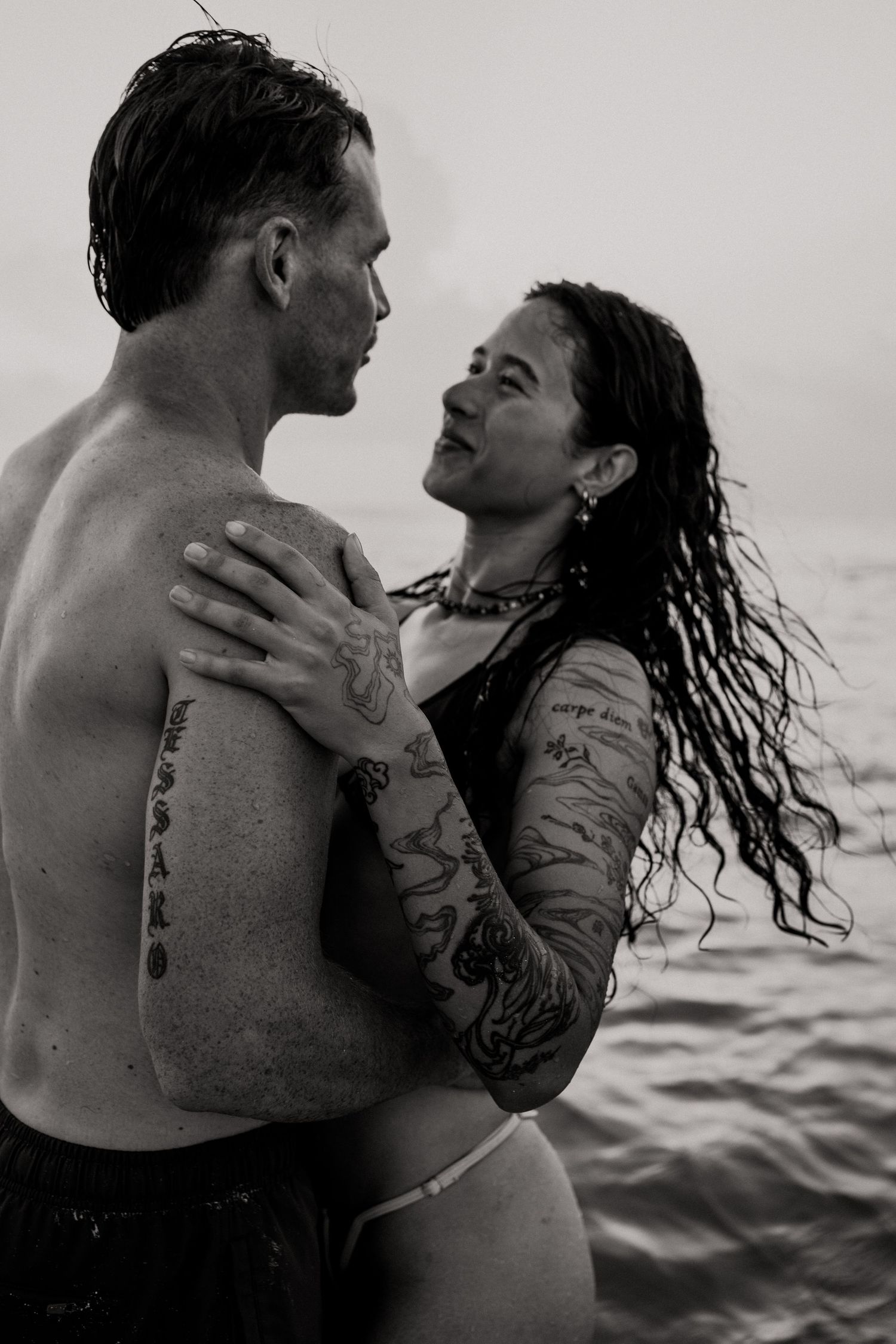 Black and white photo of a shirtless man and a tattooed woman with wet wavy hair embracing in the ocean while gazing at each other.