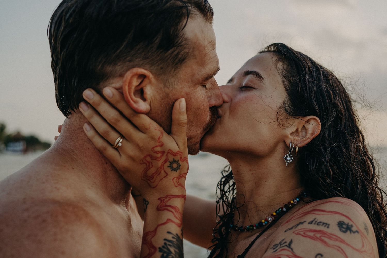 Tattooed couple with wet hair share a romantic kiss at the beach, her hand gently touching his face at sunset.