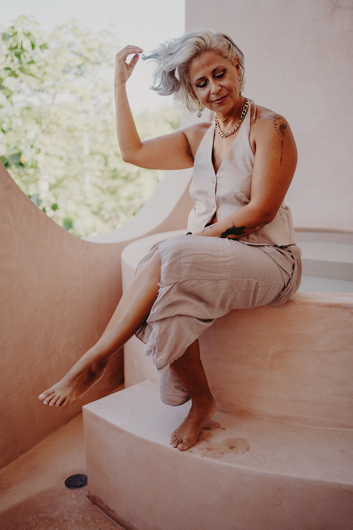 Mature woman with gray hair sits barefoot on pink steps in a linen dress, posing gracefully outdoors.