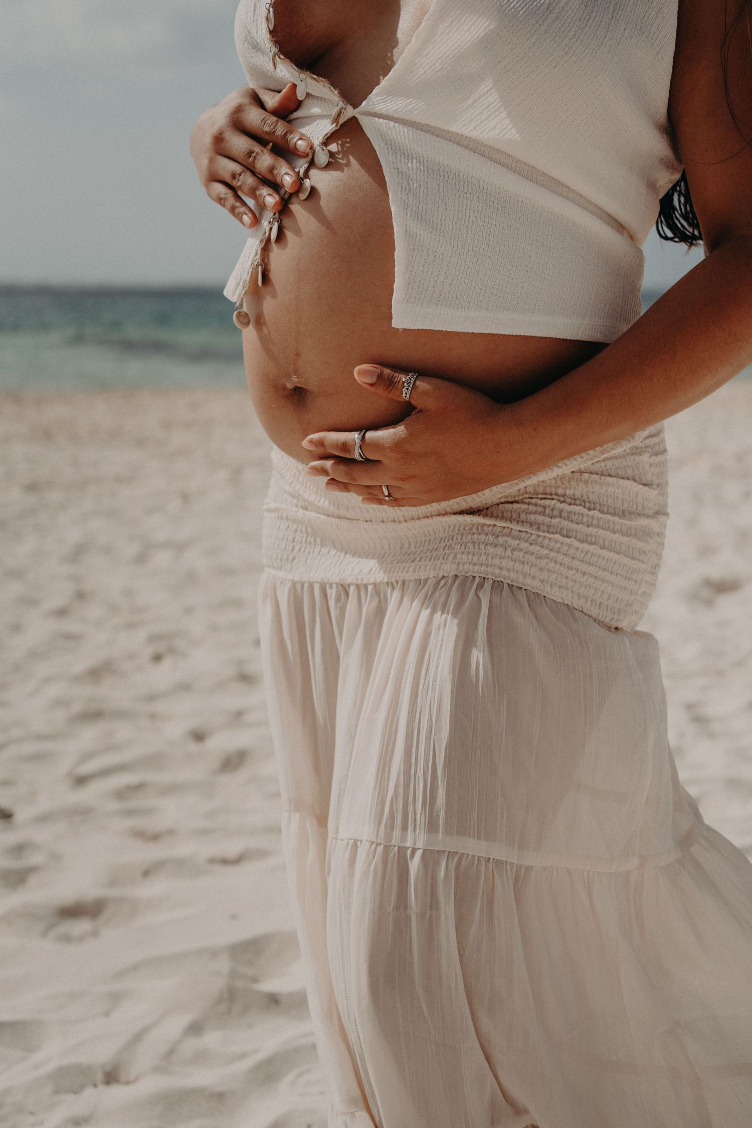 Pregnant woman in white boho crop top and maxi skirt cradling baby bump on sandy beach.