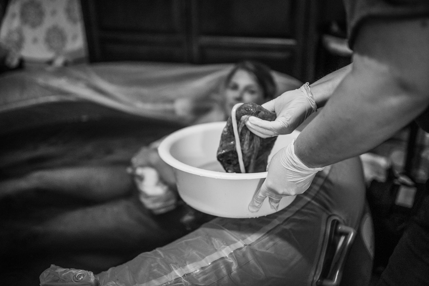 Black and white photo of a newborn baby being washed in a bowl by a gloved healthcare worker during childbirth.