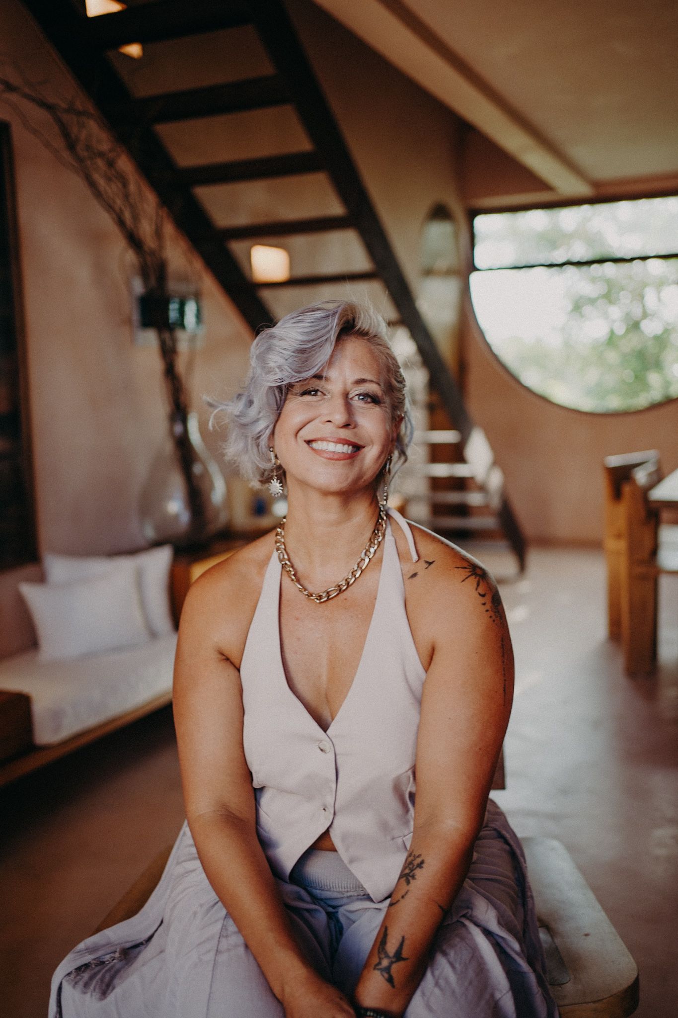 Smiling woman with silver hair, tattoos, and gold necklace sits in a stylish rustic interior with stairs and natural light.