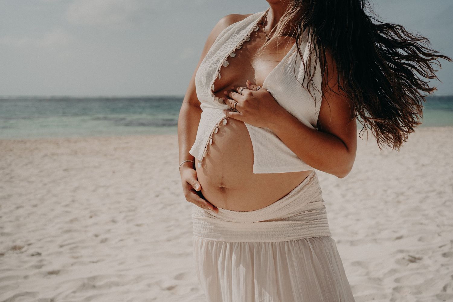 Pregnant woman in white outfit cradling her baby bump on a sandy beach with turquoise ocean in background.